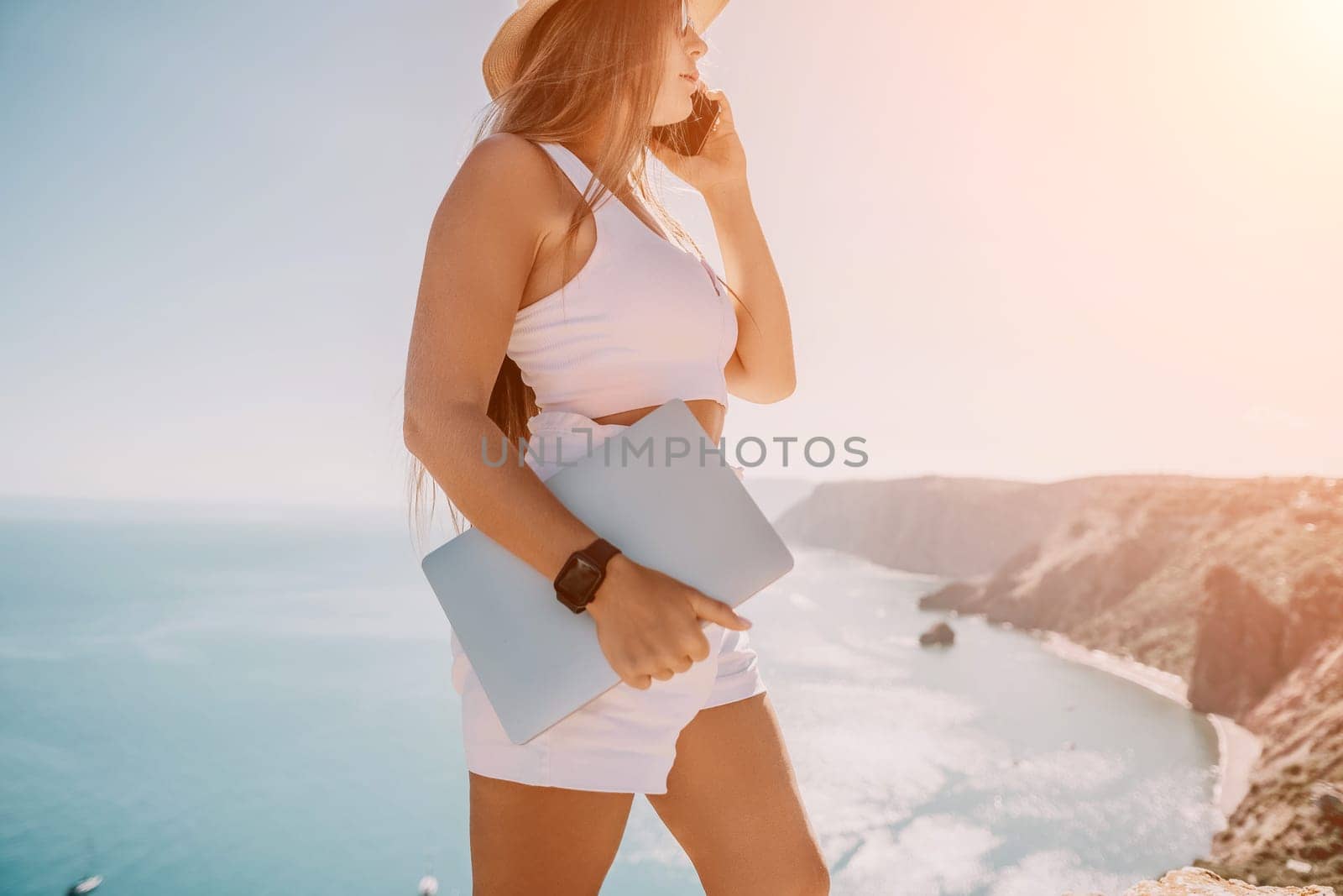 Successful business woman in yellow hat working on laptop by the sea. Pretty lady typing on computer at summer day outdoors. Freelance, travel and holidays concept.