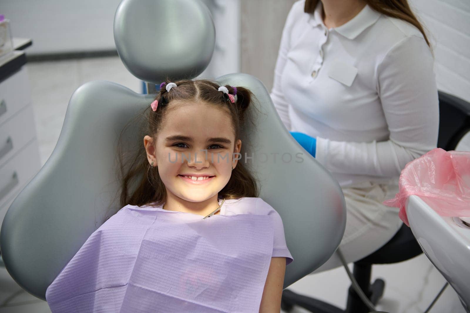 Little child girl smiles looking at camera, sitting in dentist's chair, receiving dental treatment in dentistry clinic by artgf