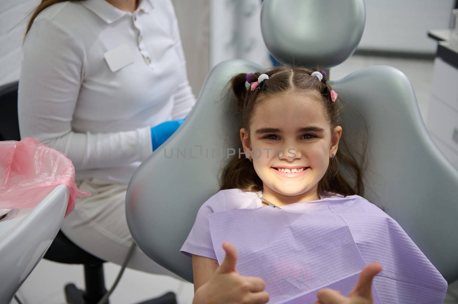 Smiling little girl at dentist appointment, sitting on dental chair, showing thumbs up after preventive dental check up in modern children's dental clinic. Pediatric dentistry. Prevention of caries