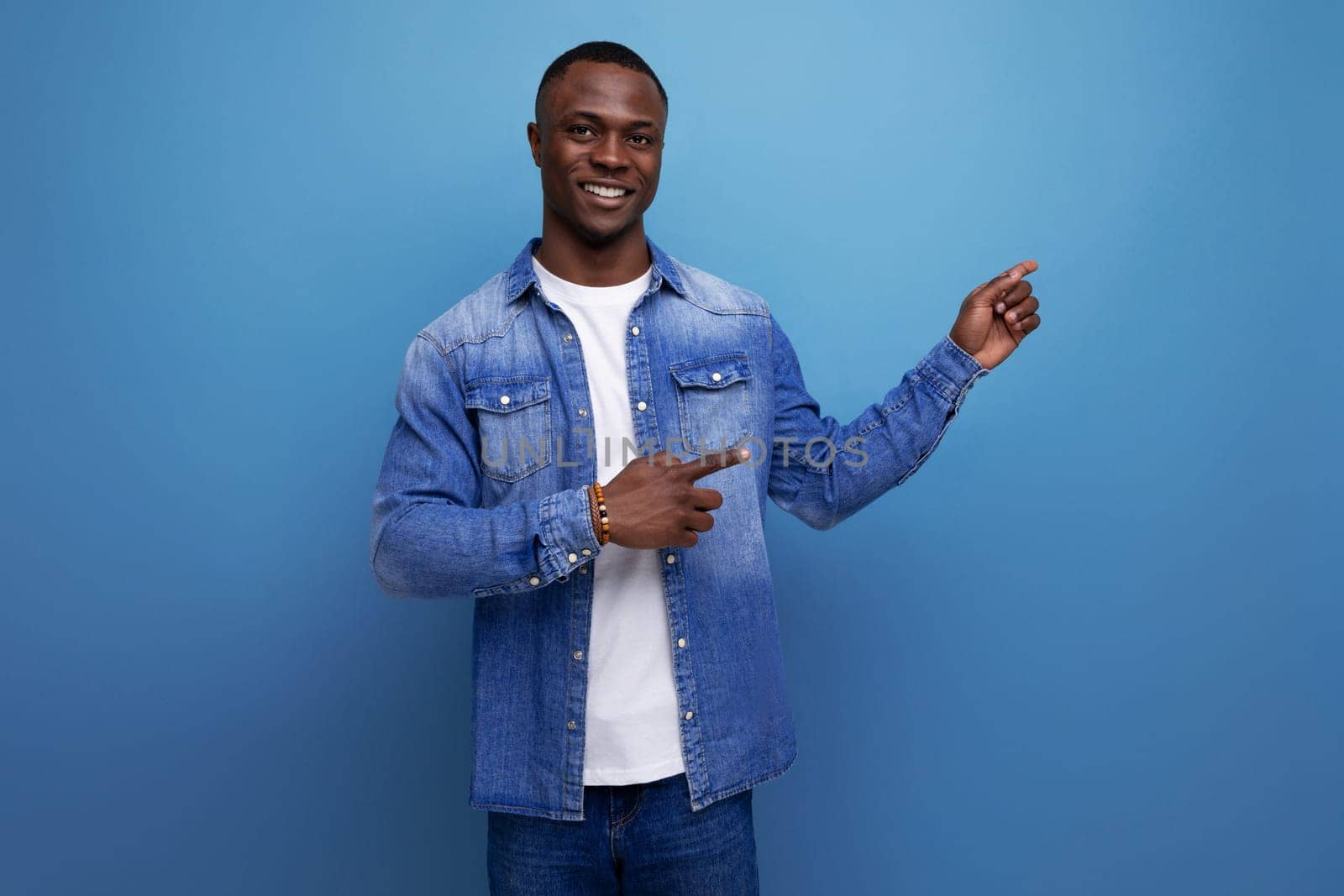 handsome young american man in denim jacket showing hand to side on blue background with copy space.