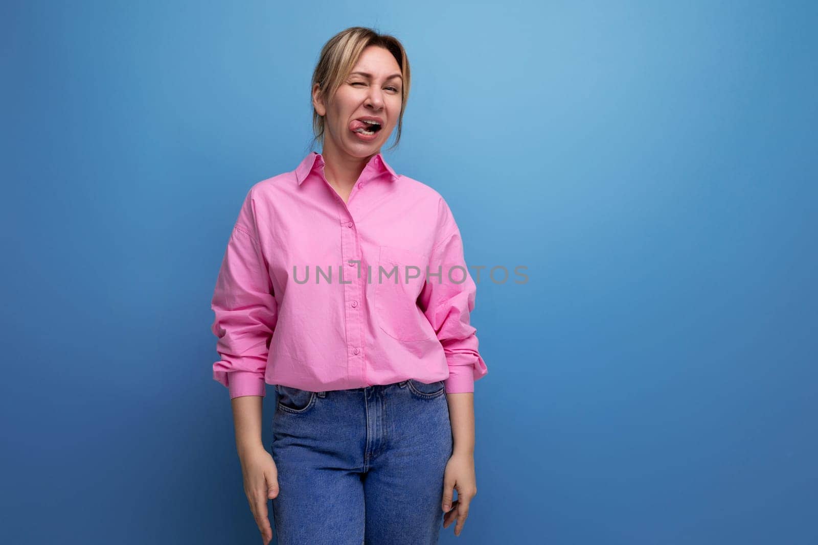 young energetic European blond leader woman with ponytail dressed in a pink blouse makes a grimace against the background with copy space.