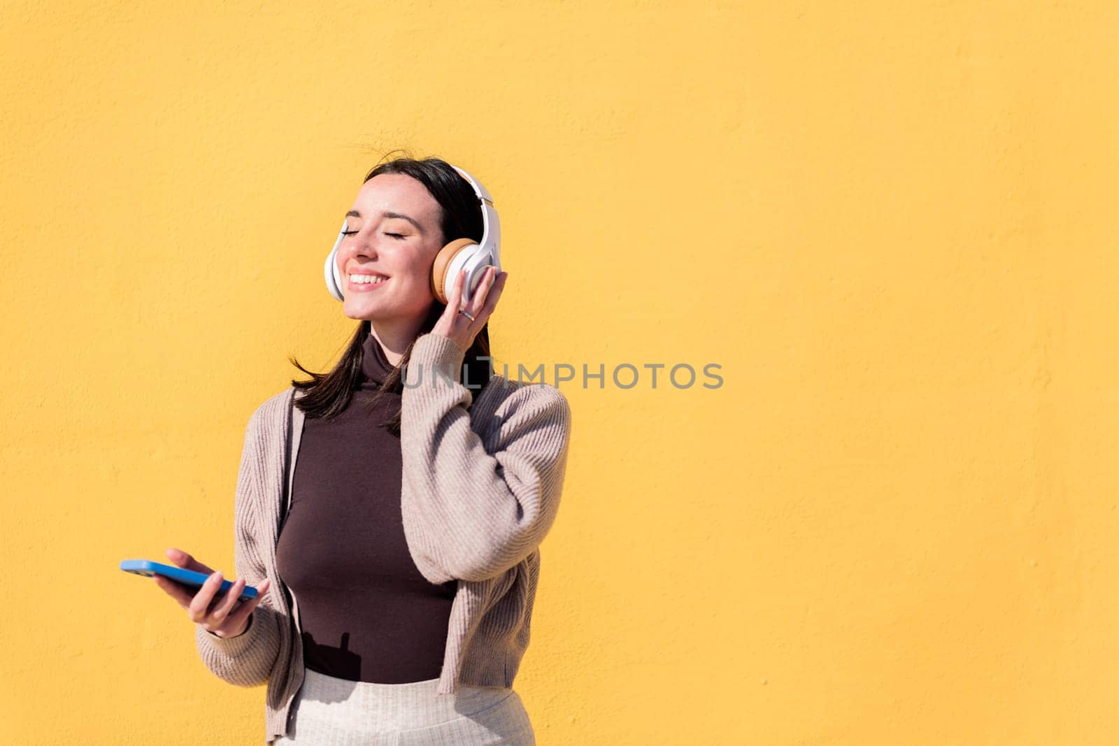woman enjoying happy listening to music from mobile phone in headphones with a yellow wall in the background, concept of rhythm and positive people, copy space for text