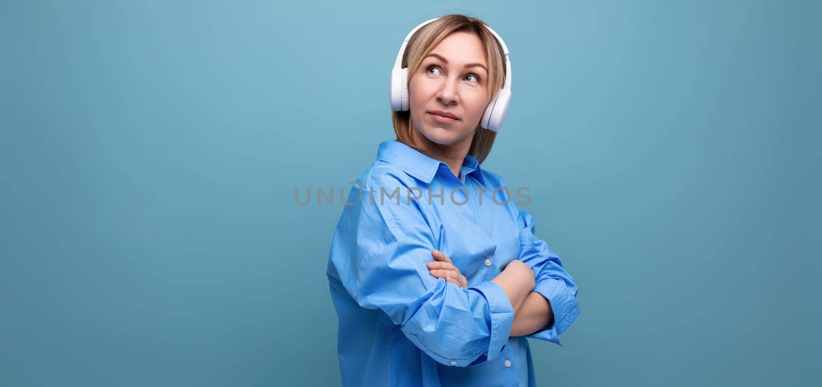 widescreen photo of a charming european girl in a casual shirt listening to music in big white headphones on a blue isolated background by TRMK