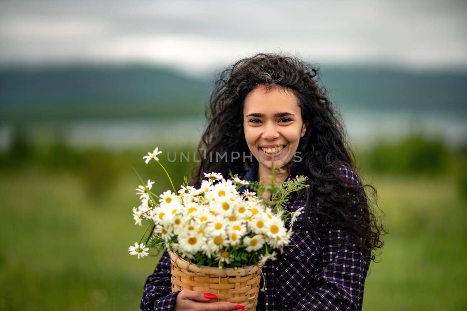 A woman stands on a green field and holds a basket with a large bouquet of daisies in her hands. In the background are mountains and a lake