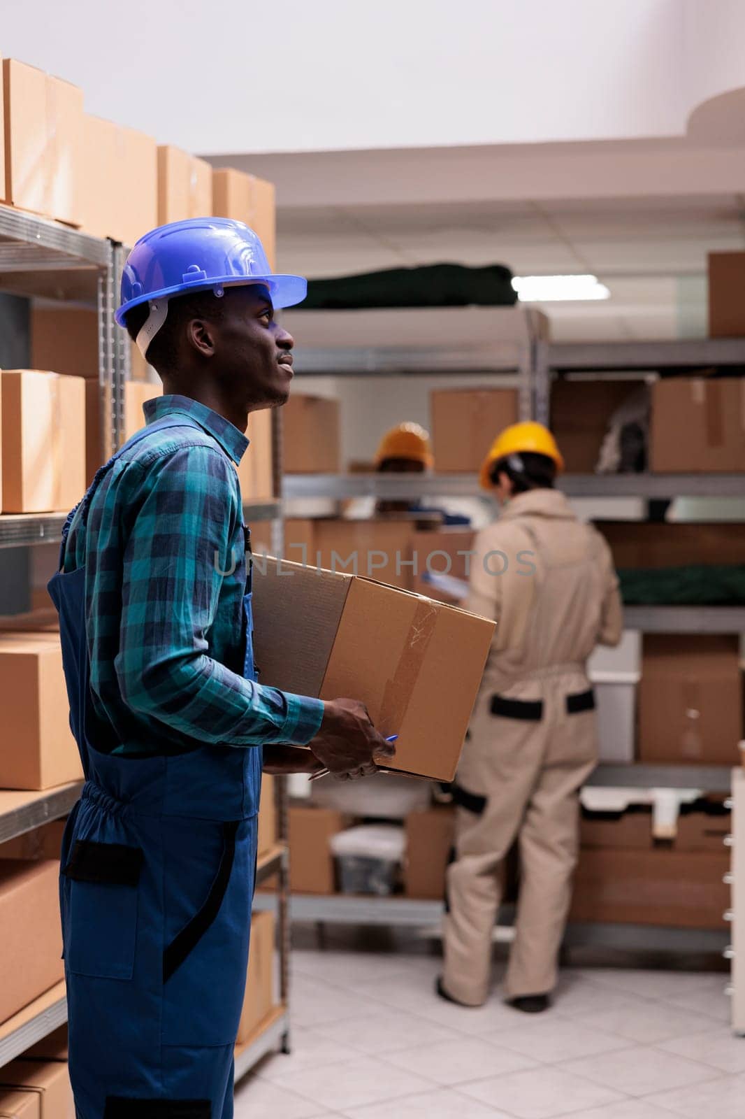 Warehouse manager wearing protective helmet carrying parcel and making stock supply inventory. Young african american storehouse operator in uniform monitoring packages maintenance
