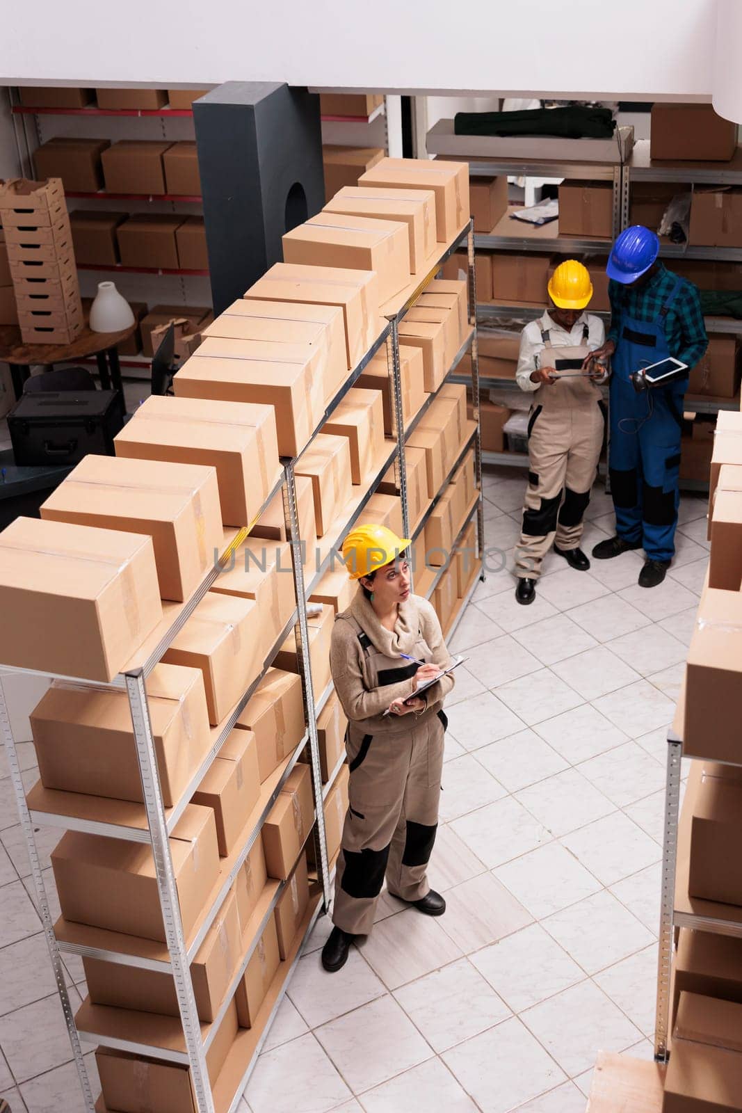 Warehouse assistants standing near cardboard boxes shelves and doing inventory. Caucasian and african american coworkers team wearing protective helmets working in delivery service storage
