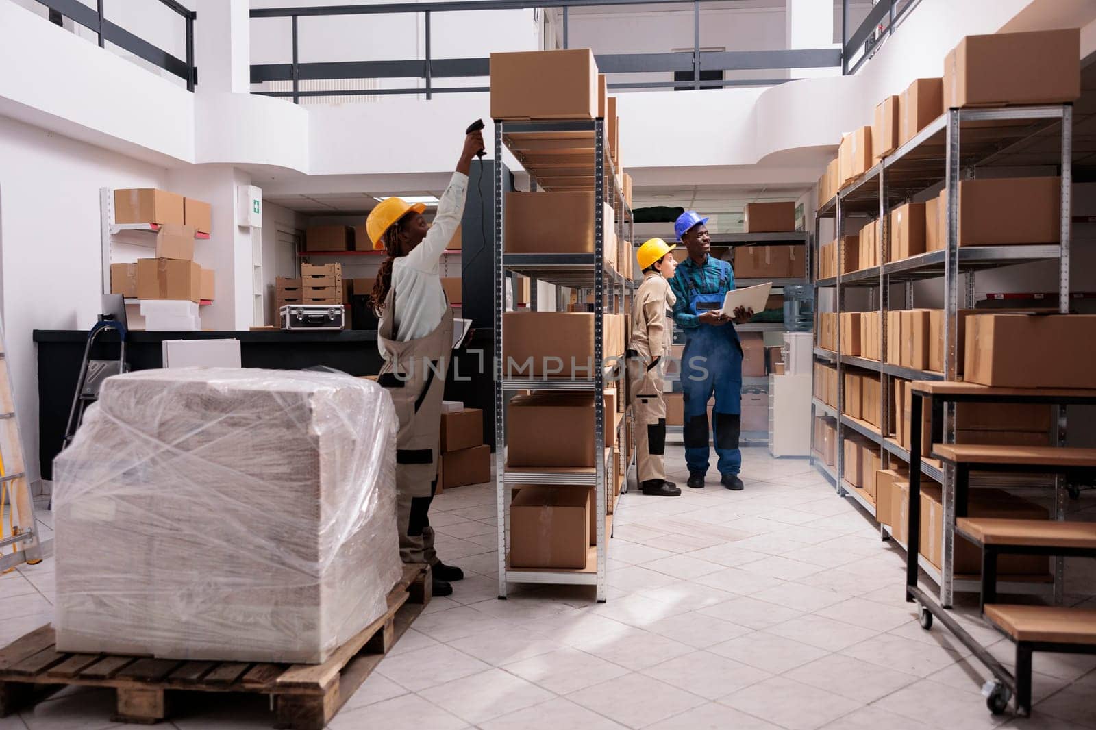 Industrial storehouse employees scanning cardboard boxes barcodes and checking stock supply on laptop. Diverse warehouse coworkers standing near parcels shelves and working