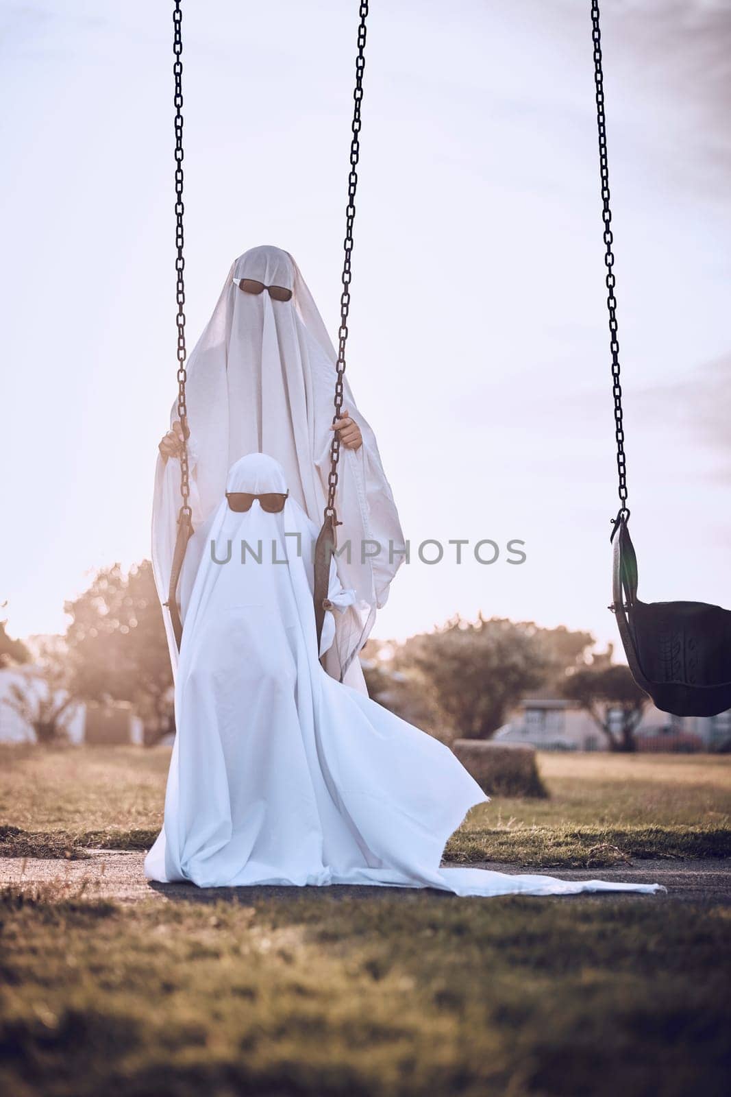 Halloween, costume and ghost couple at park, kid enjoy scary holiday with parent. Family, festival and child with mom or dad in spooky white sheet dress up with sunglasses sitting on playground swing by YuriArcurs