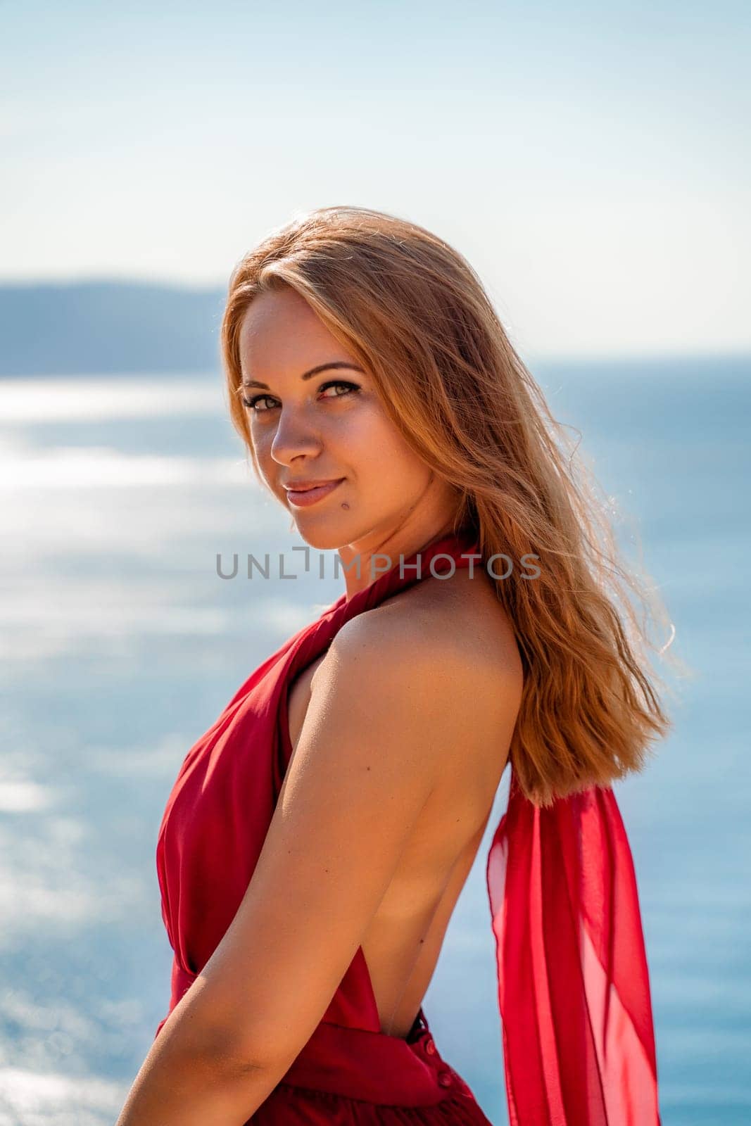 Smiling young woman in a red dress looks at the camera. A beautiful tanned girl enjoys her summer holidays at the sea. Portrait of a stylish carefree woman laughing at the ocean. by Matiunina