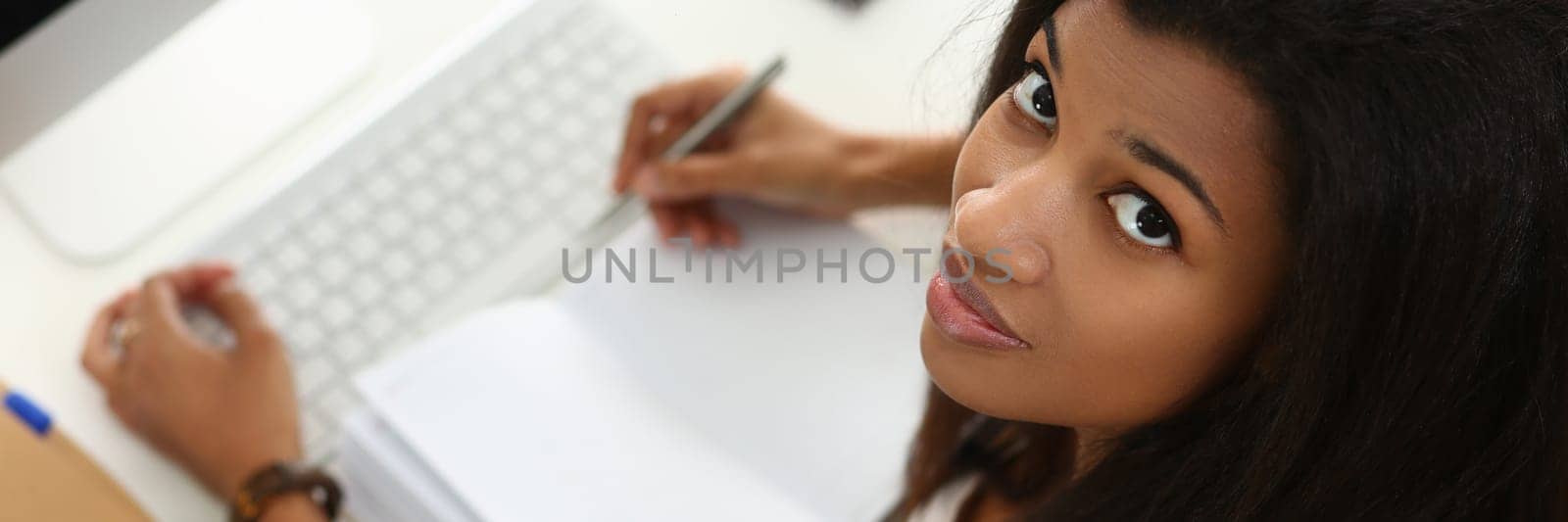Smiling afro woman sitting at table holding pen and notebook in home office by kuprevich
