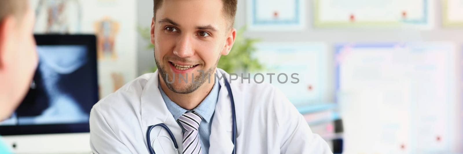 Young smiling male doctor in medical uniform takes patient. Smiling and chatting in medical clinic