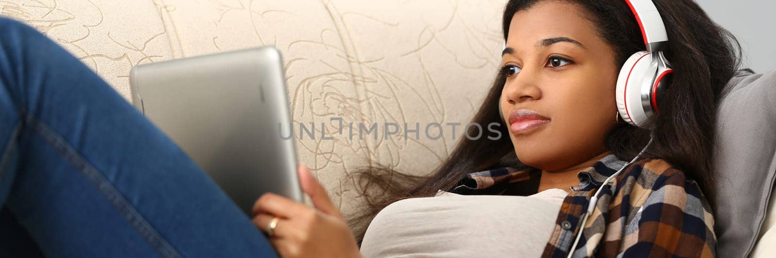 Happy african american young woman lying on sofa with tablet computer and headphones listening to music at home by kuprevich