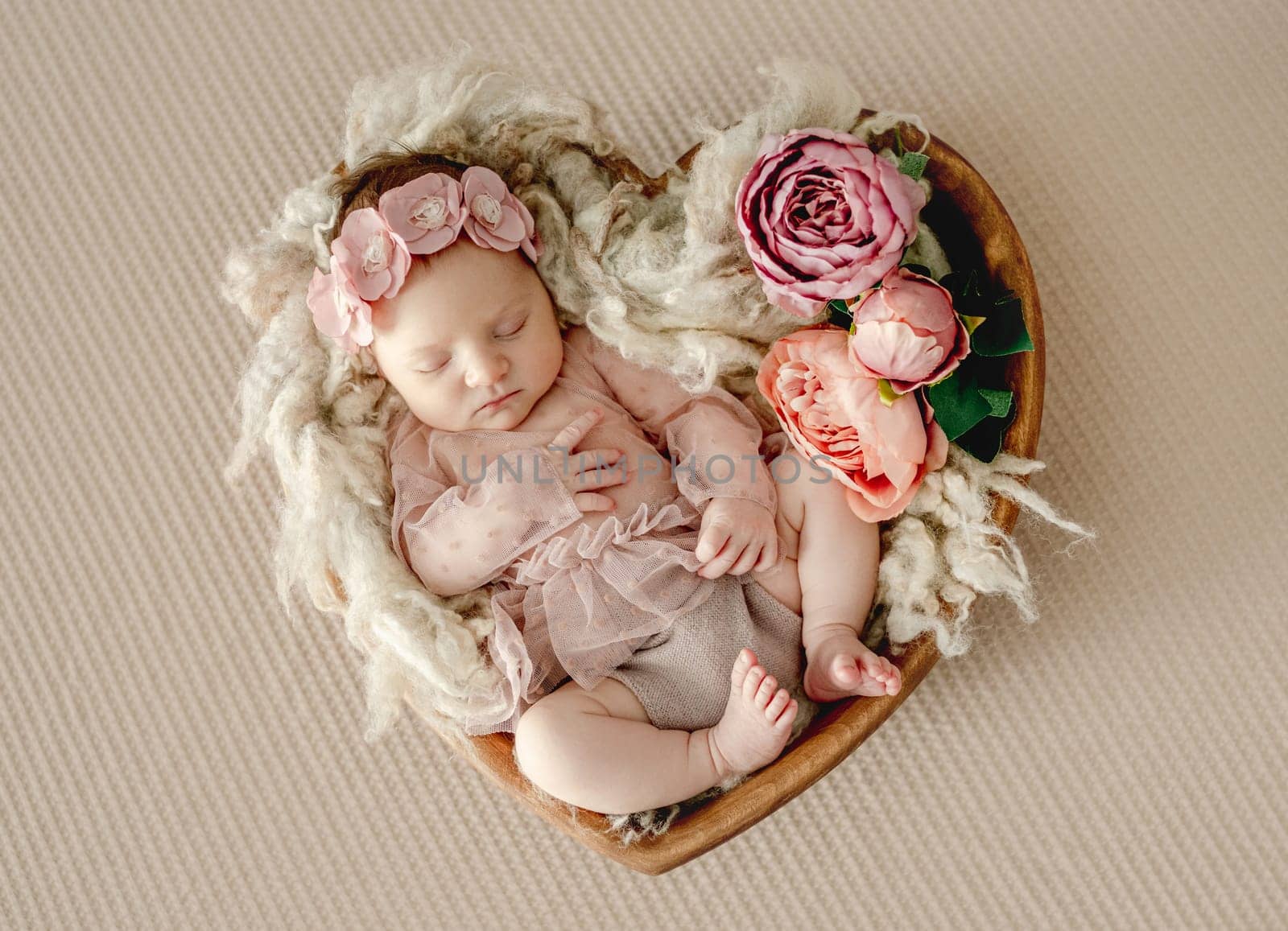 Newborn baby girl wearing dress sleeping in heart shape basket with toy. Cute infant child kid in wreath napping on fur