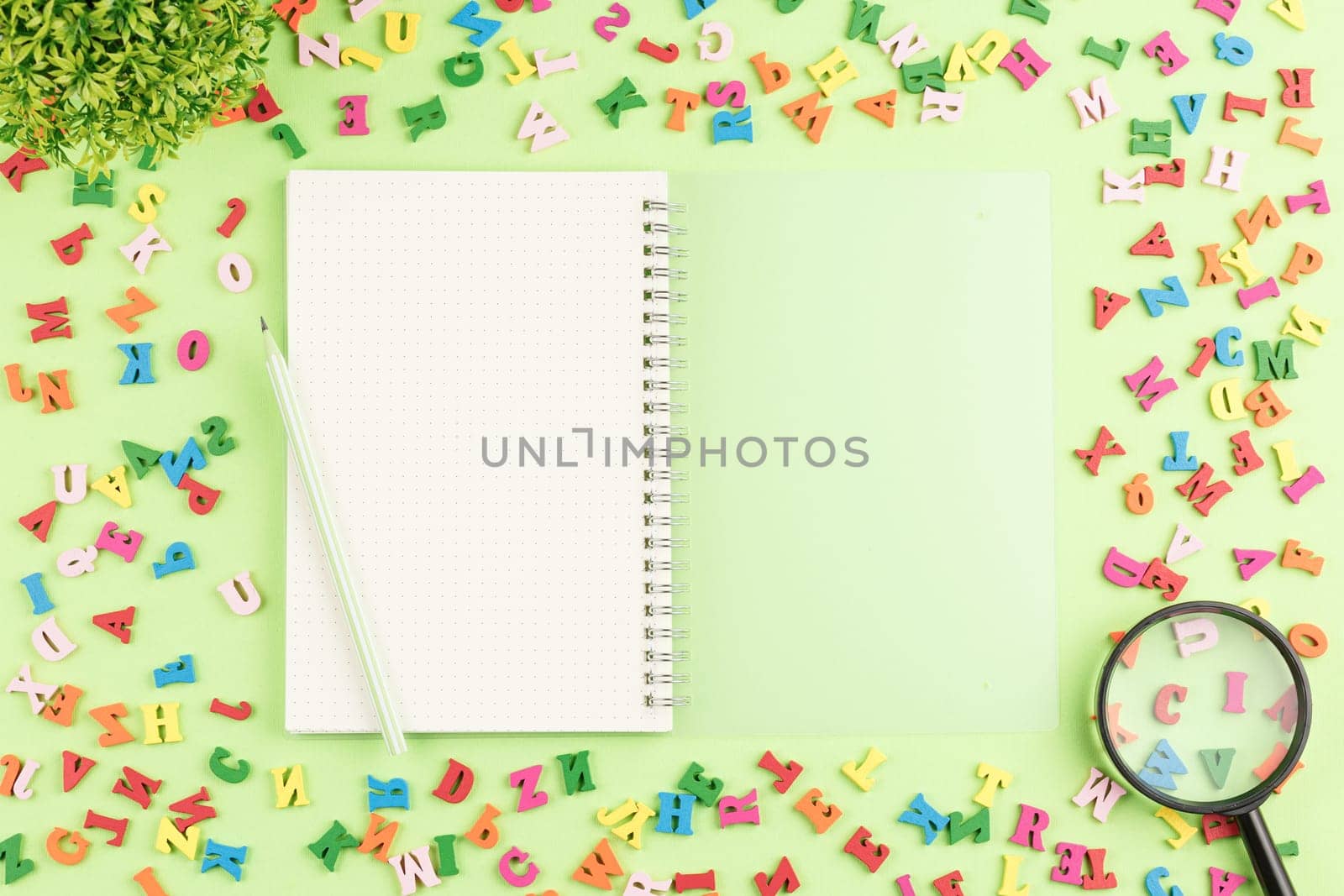 Open notebook with a pencil and a magnifying glass on a green background. Top view. Colored wooden alphabet on a school desk. Flat lay. Back to school concept.
