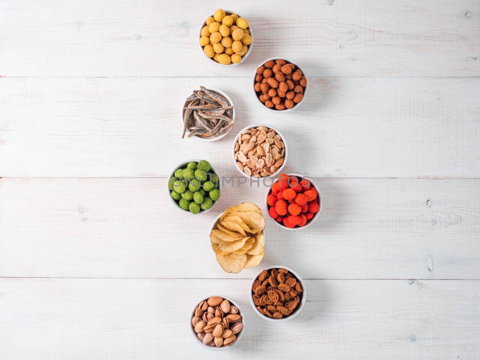 Assortment of different snack for beer, wine, party. Peanuts in coconut glaze, green vasabi, red spicy chilli, yellow cheese glaze, chips, pistachio, crackers, fish on white wooden table. Copy space