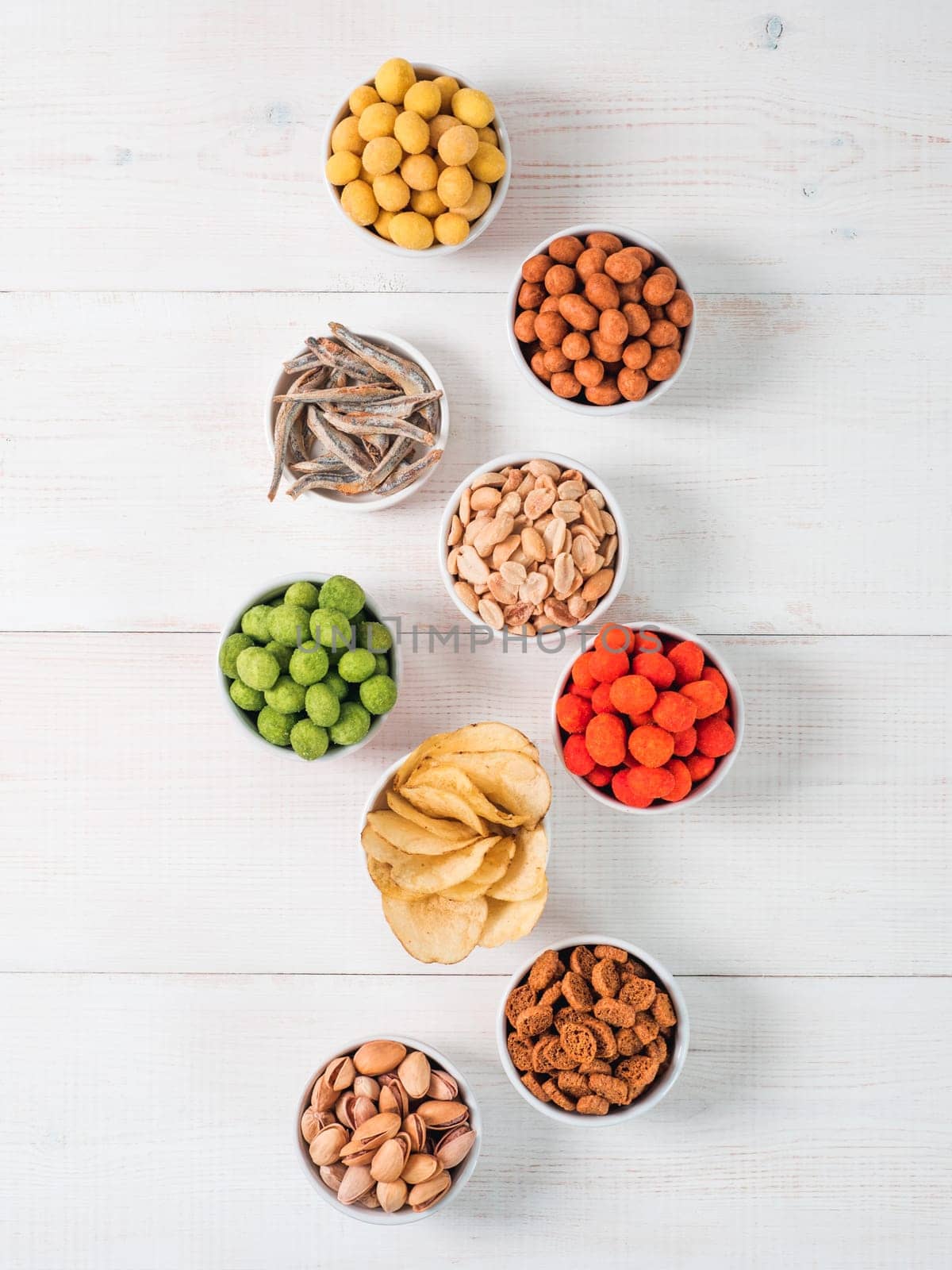 Assortment of different snack for beer,wine,party.Peanuts in coconut glaze,green vasabi,red spicy chilli,yellow cheese glaze and chips,pistachio,crackers,fish on white wooden table.Copyspace.Vertical.