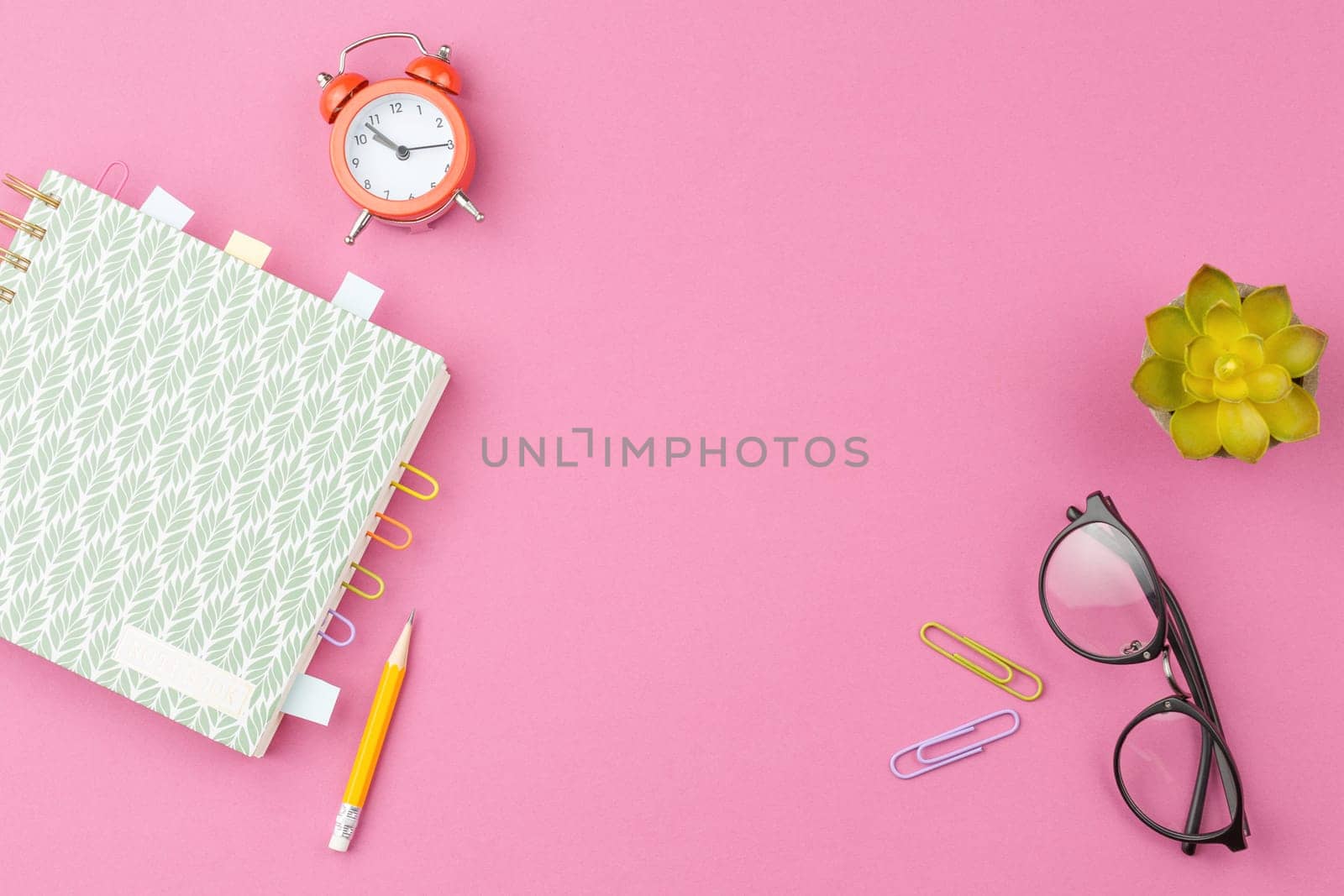 Spiral notebook with foliage ornament, bookmarks from paper clips and sheets for notes, glasses, pencil, alarm clock and a flower in a pot on a pink background. Top view.