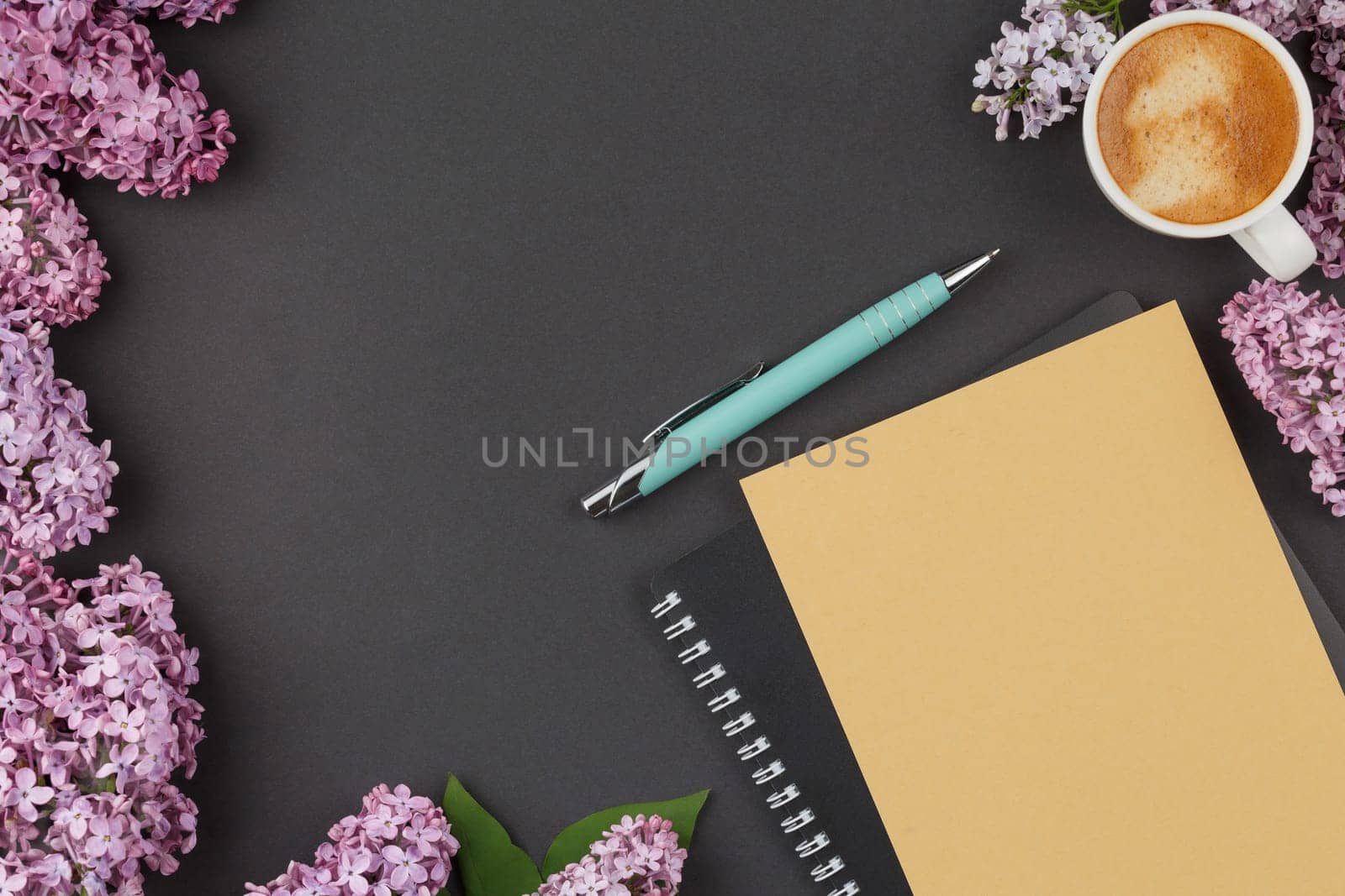 Black spiral notebook with pen, coffee cup and lilac flowers on an isolated background. Top view.