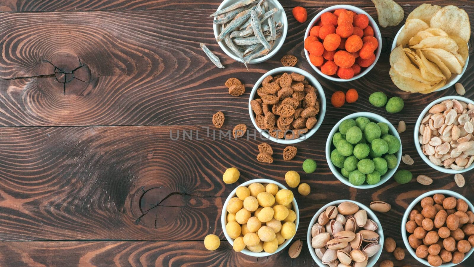 Assortment of different snack for beer, wine, party. Peanuts in coconut glaze, green vasabi, red spicy chilli, yellow cheese glaze, chips, pistachio, crackers, fish on brown wooden table. Copy space