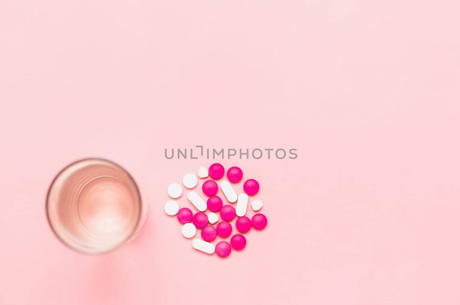 Top view of a glass of drinking water with piles of medicine pills on pink background for healthcare concept.