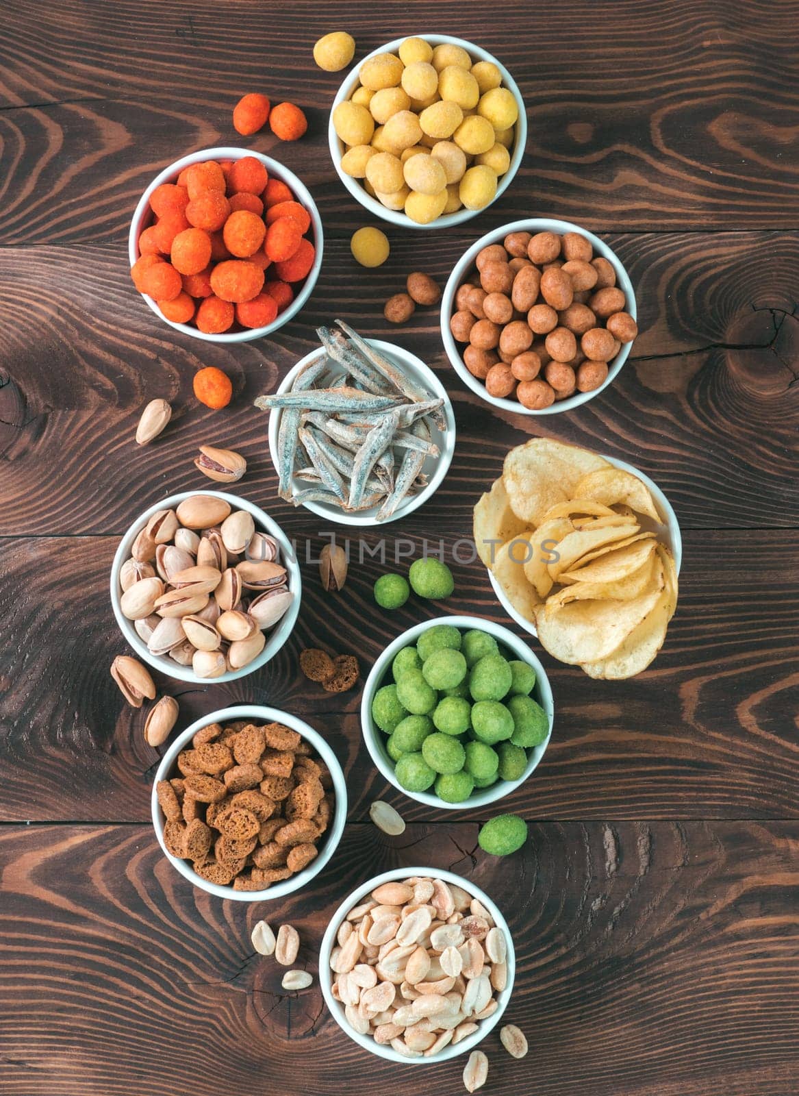 Assortment of different snack for beer, wine, party. Peanuts in coconut glaze, green vasabi, red spicy chilli, yellow cheese glaze, chips, pistachio, crackers, fish on brown wooden table. Copy space