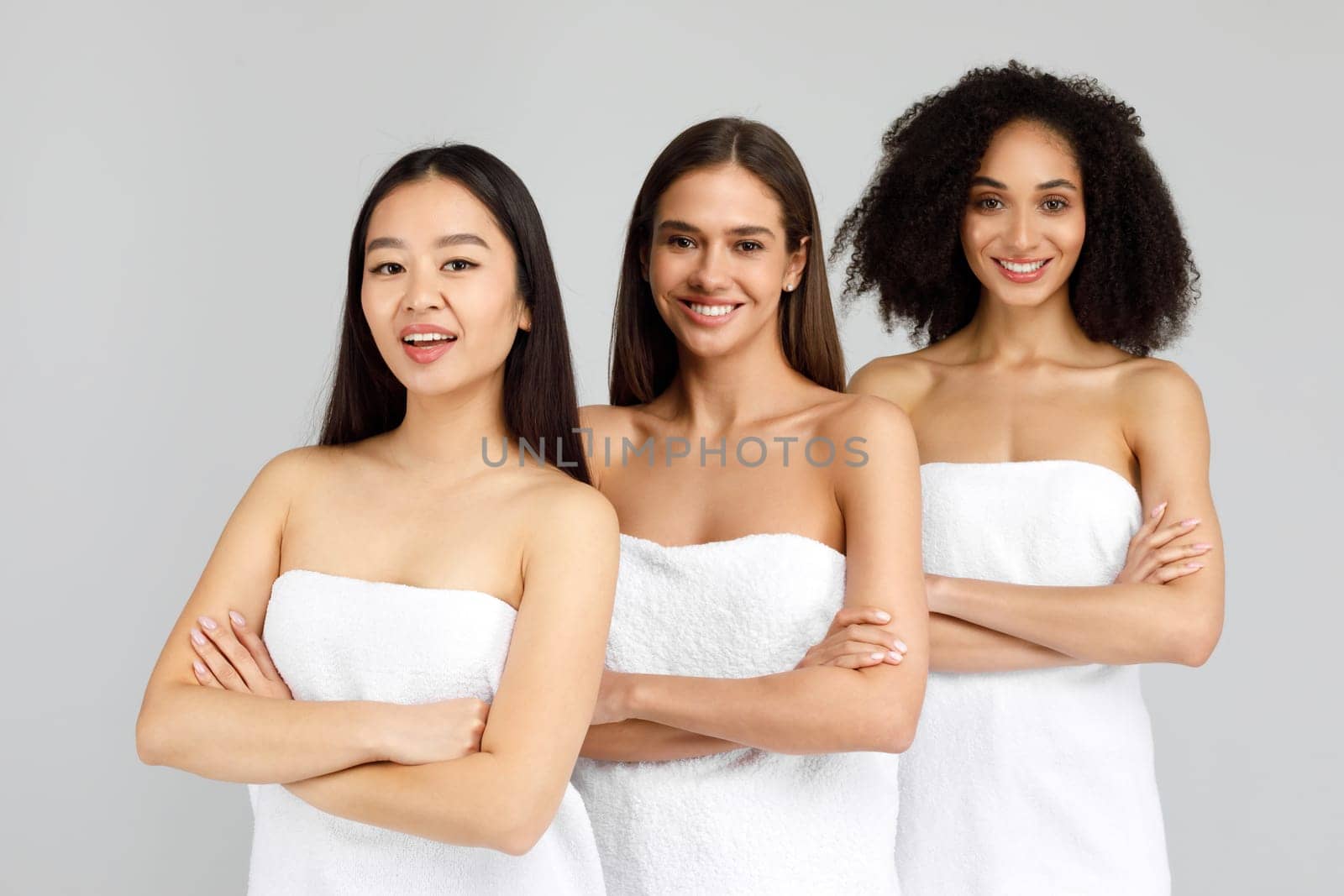 Portrait of three young beautiful multiracial ladies posing wrapped in towels with folded arms, smiling at camera while standing in line over grey background