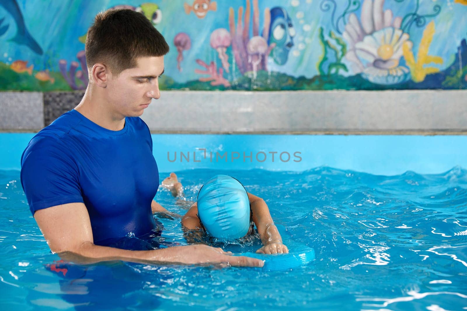 Trainer teaching little boy how to swim in indoor pool with pool floating board by Mariakray