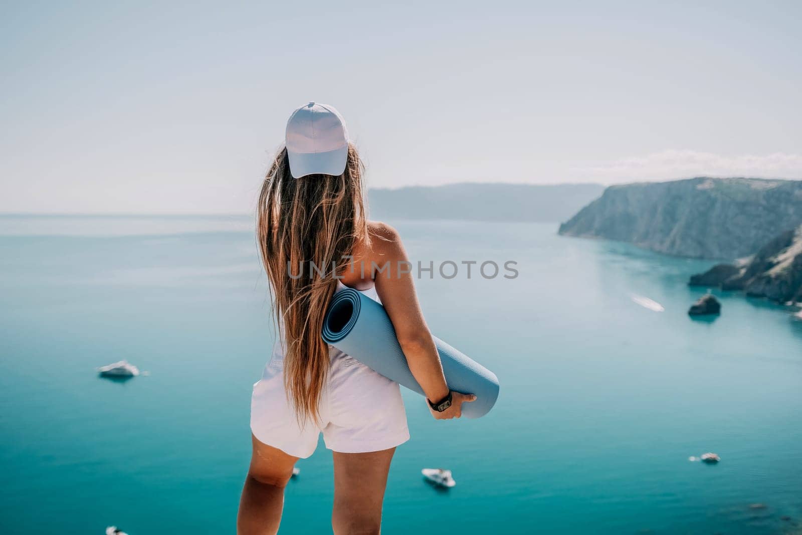 Woman with yoga mat over calm azure sea. Well looking middle aged woman with long hair, fitness instructor with blue yoga mat near the sea. Female fitness yoga routine concept. Healthy lifestyle. by panophotograph