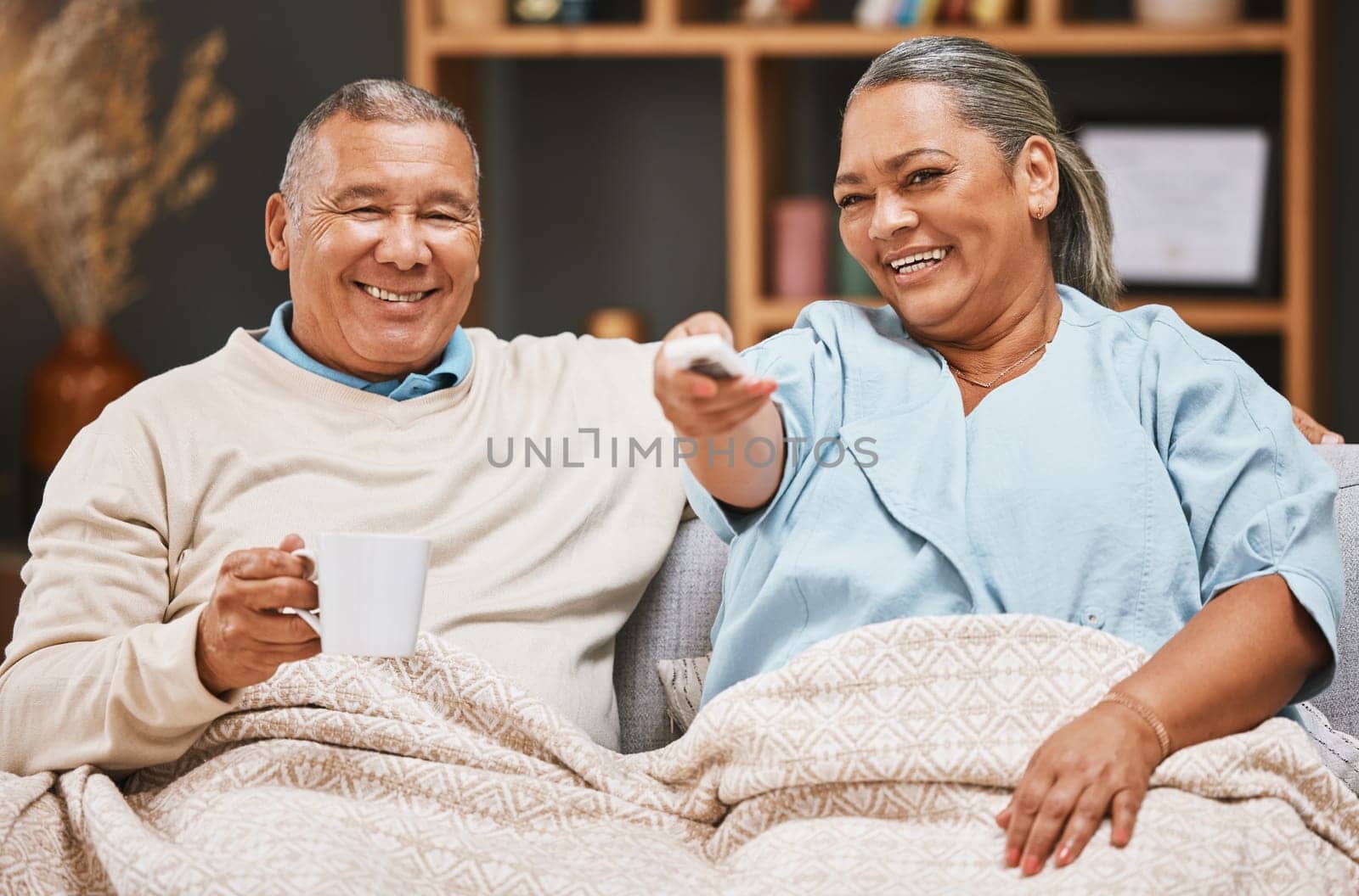 Coffee, relax and old couple watching tv with blanket on sofa in home living room laughing at comic film. Retirement portrait, valentines love and happy elderly man and woman streaming on television