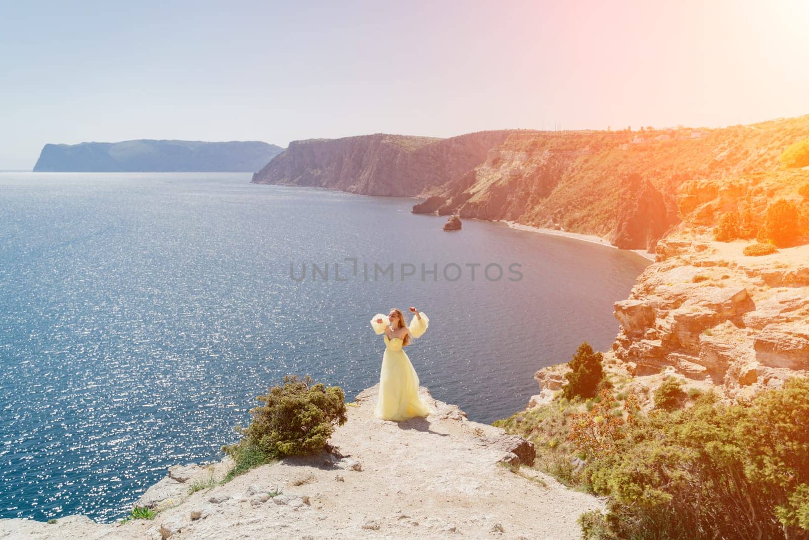 Woman yellow dress sea. Side view Young beautiful sensual woman in yellow long dress posing on a rock high above the sea at sunset. Girl in nature against the blue sky by Matiunina