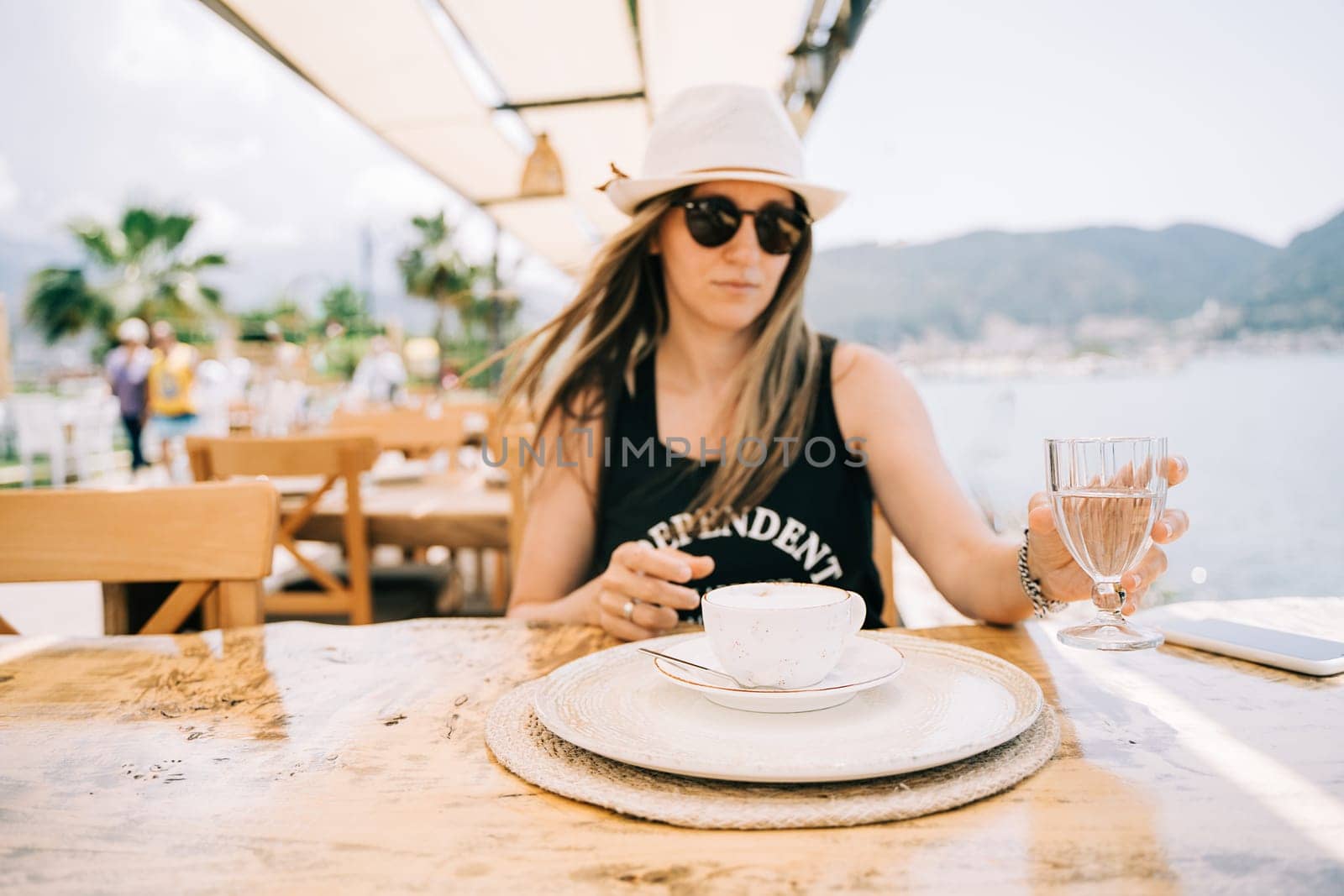Young woman female in sunglasses and hat drinking coffee in a street outdoors seaside cafe restaurant coffee shop with scenery sea and mountains in the background. Hello summer holiday vacation.
