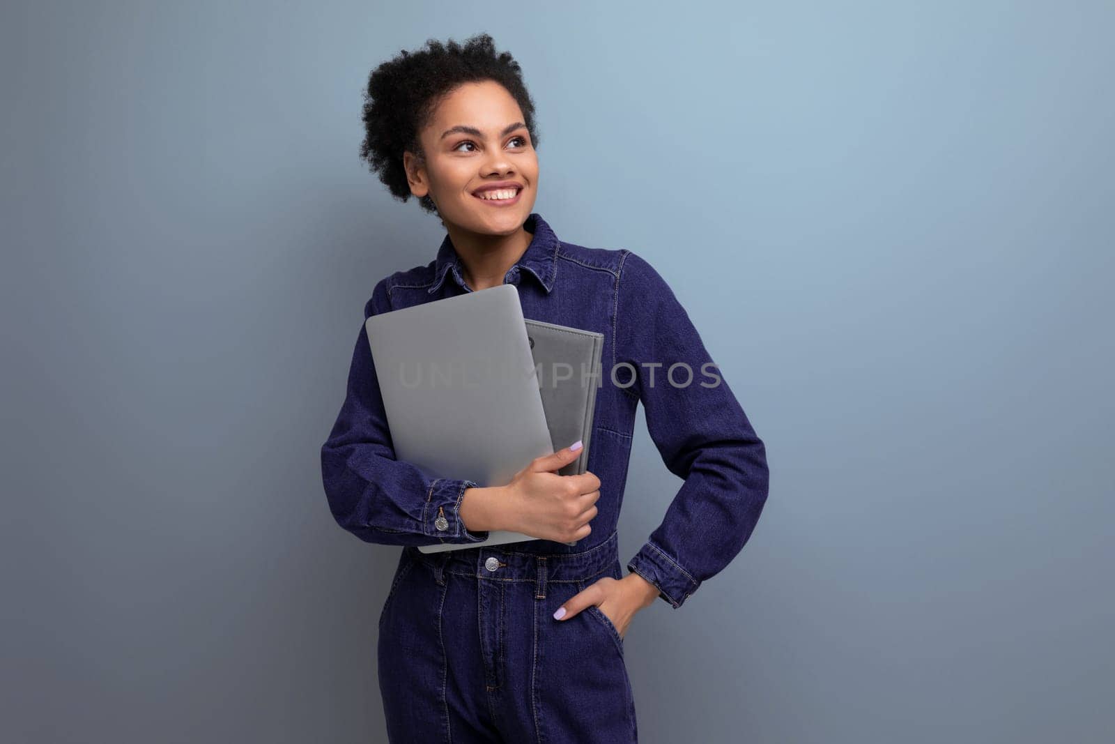 a slender young hispanic brunette woman with fluffy curly hair in a blue denim suit will hold documents and a laptop for work by TRMK