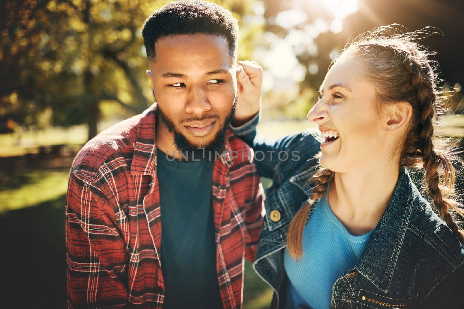 Interracial couple, laughing and smile in nature for playful love, bonding and funny relationship. Happy woman holding man ear with laugh for fun loving care, affection or playing in a park outdoors.