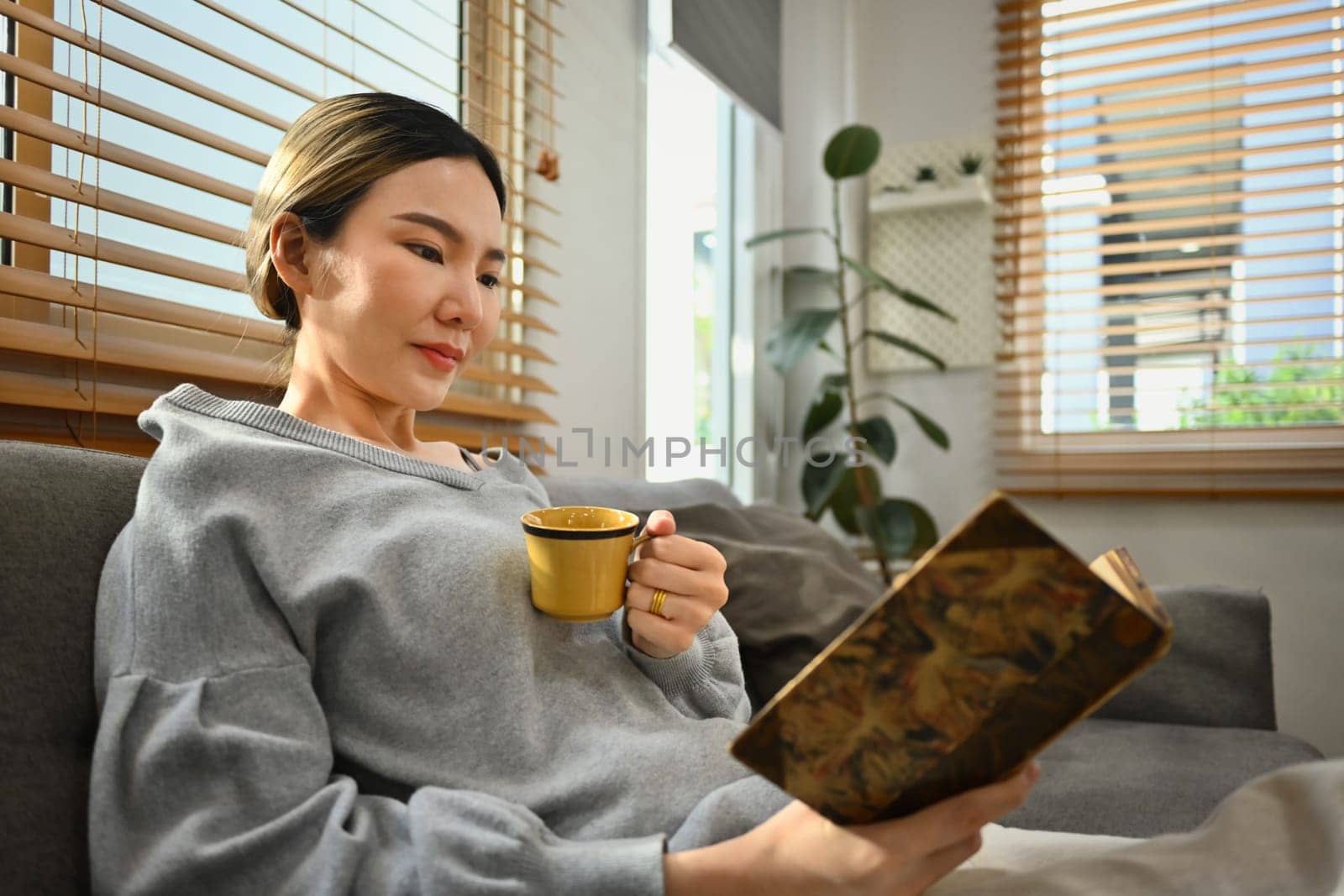 Relaxed young woman drinking hot tea and reading book on couch in living room. People, leisure and lifestyle concept.