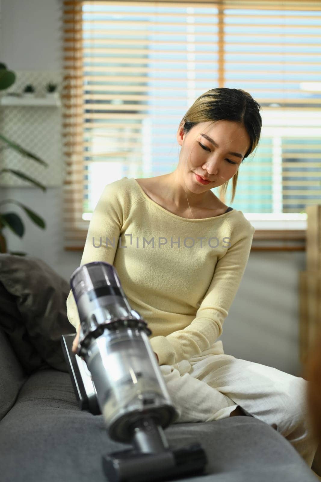 Image of asian female housewife vacuuming sofa with cordless handheld vacuum cleaner. Housework concept.
