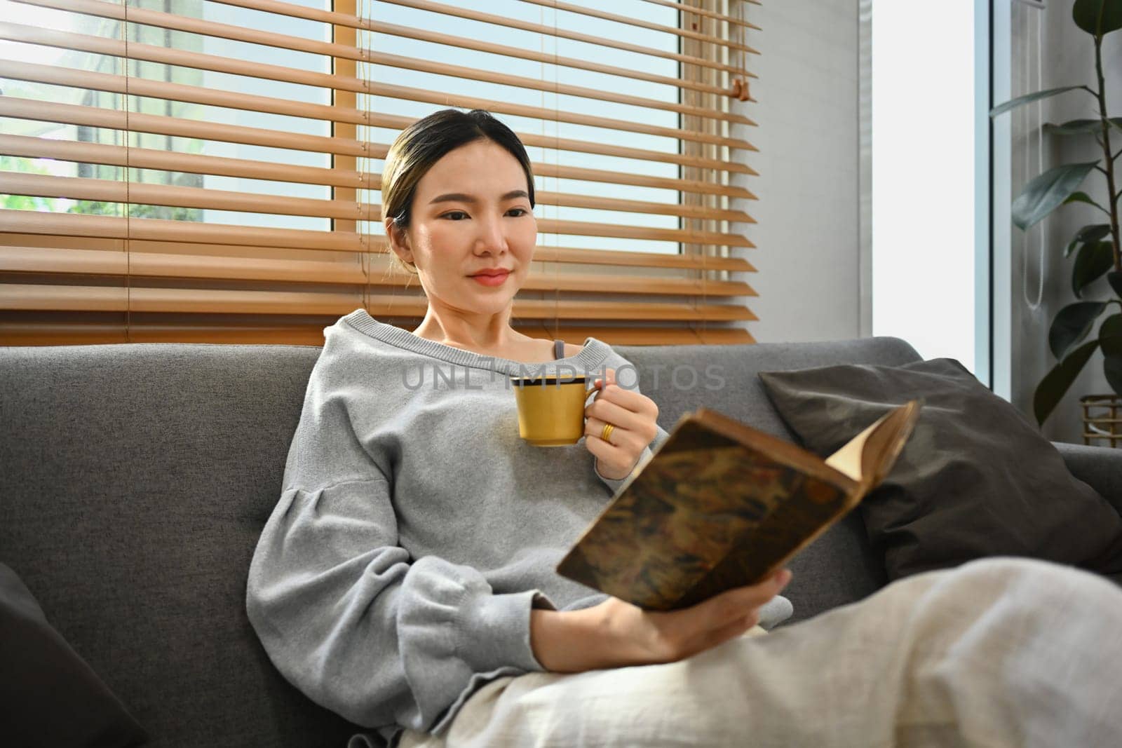 Beautiful millennial woman drinking hot tea and reading book on couch. People, leisure and lifestyle concept.