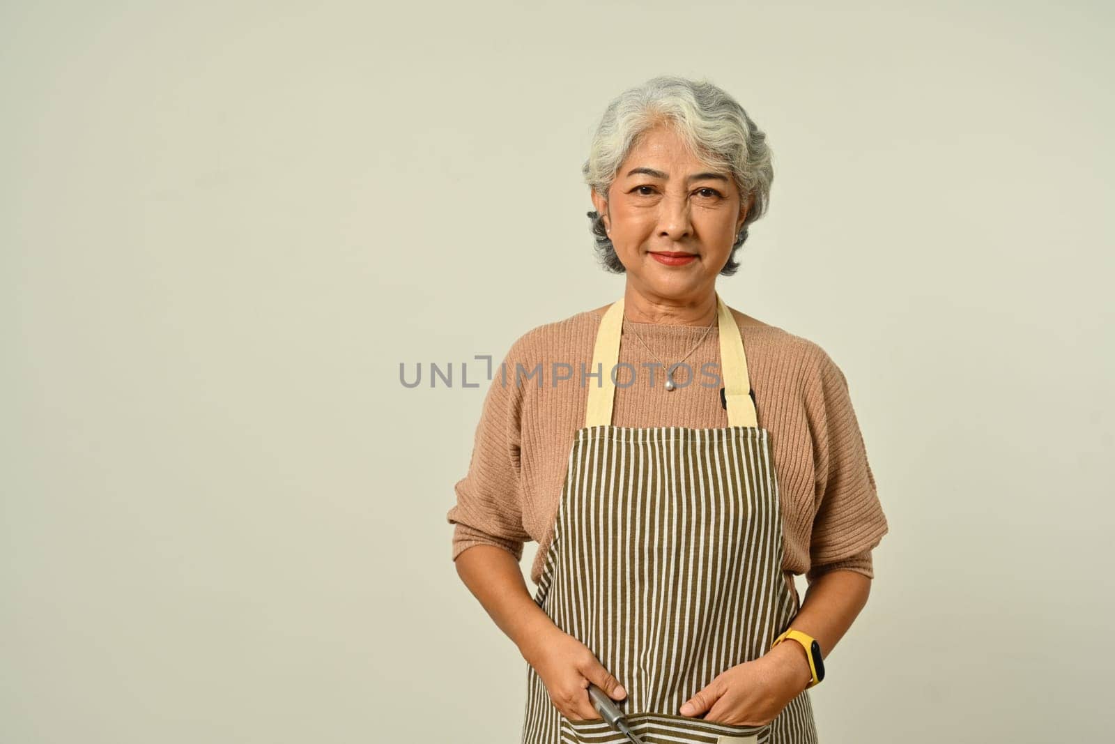 Positive grey haired woman 50s wearing apron standing isolated on gray background. People, household, small business owner concept.
