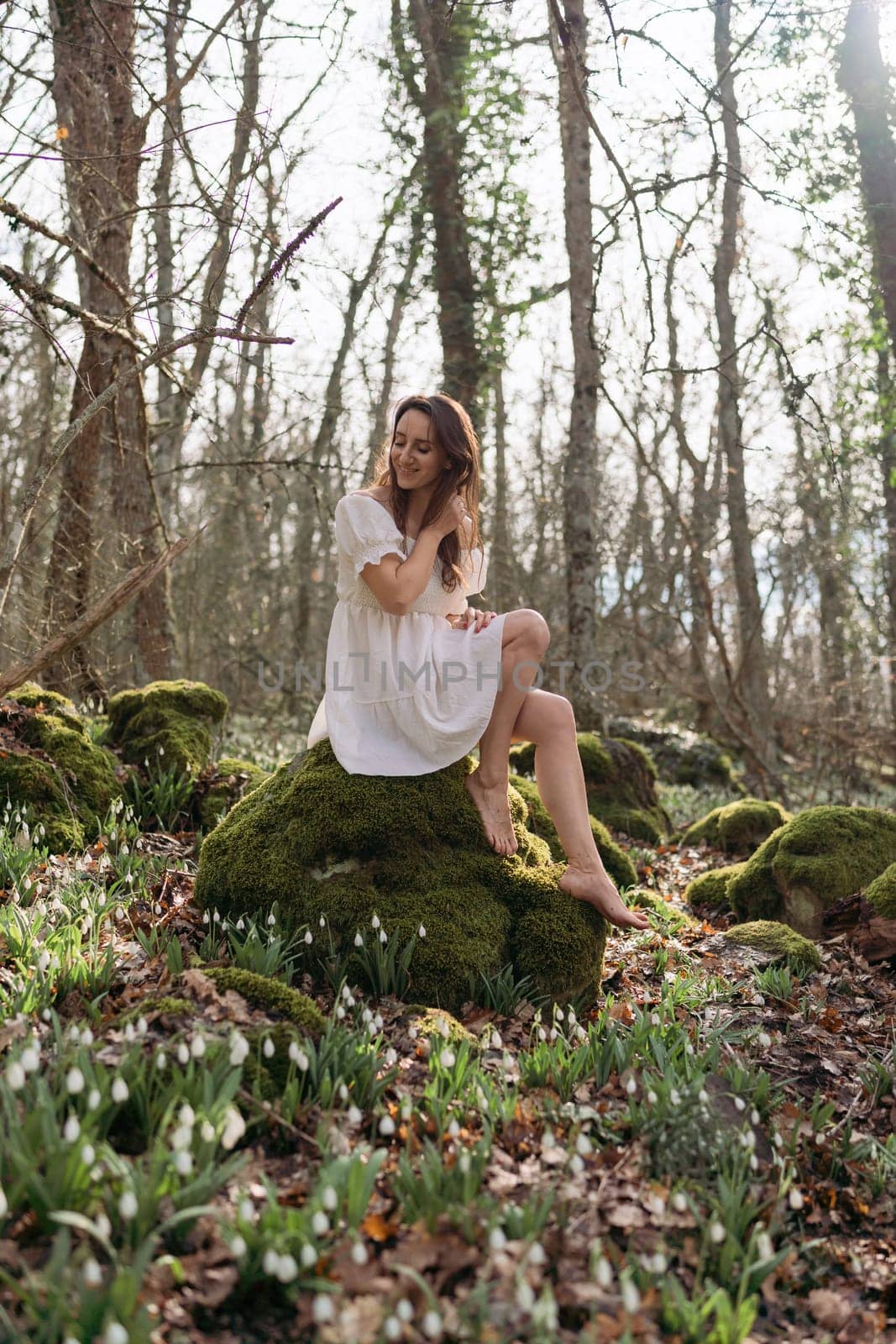 Portrait of a woman in the forest. She is sitting in a white dress on a meadow with snowdrops in a spring forest.