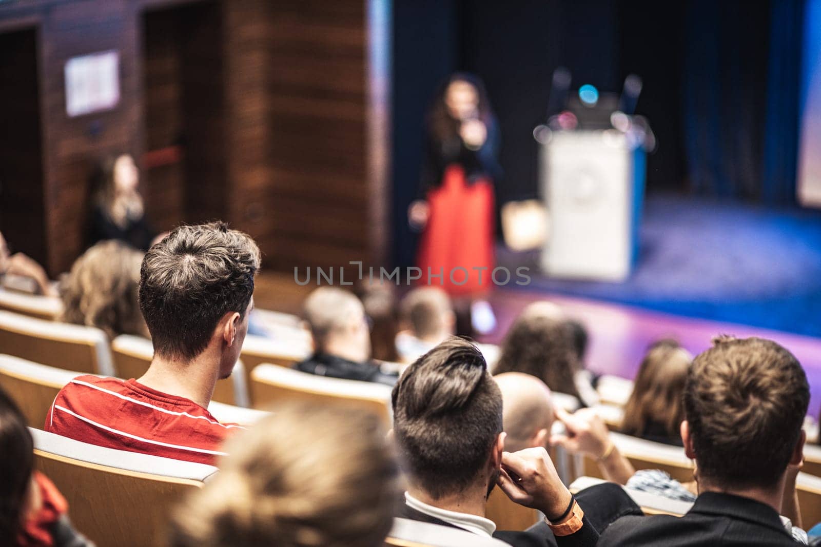 Business and entrepreneurship symposium. Female speaker giving a talk at business meeting. Audience in conference hall. Rear view of unrecognized participant in audience.