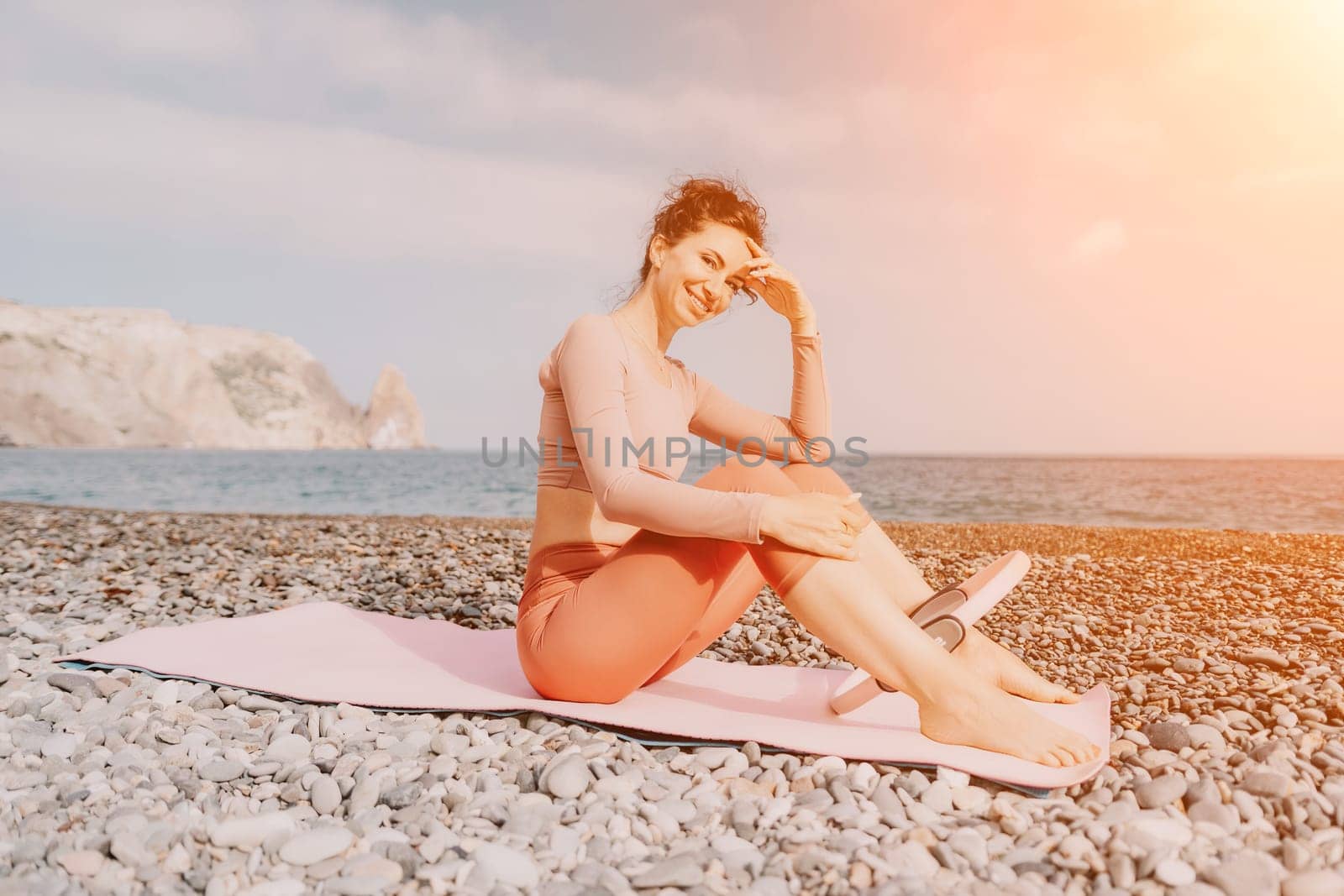Middle aged well looking woman with black hair doing Pilates with the ring on the yoga mat near the sea on the pebble beach. Female fitness yoga concept. Healthy lifestyle, harmony and meditation.