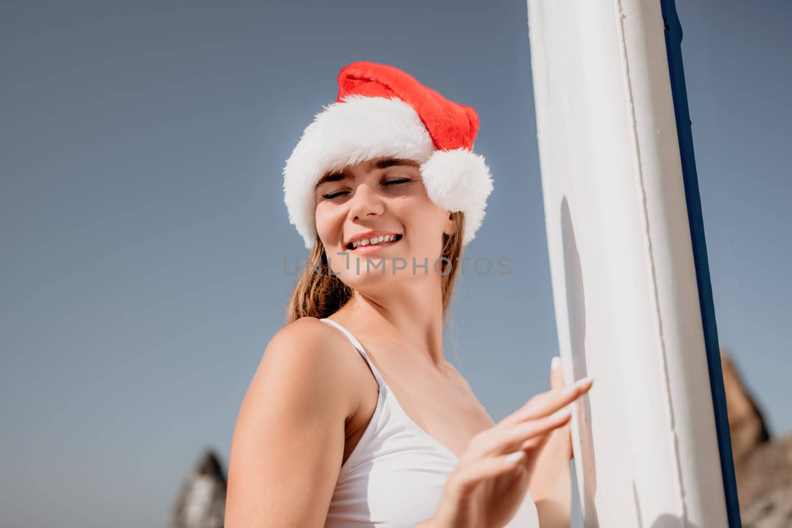 Woman sea sup. Close up portrait of happy young caucasian woman with long hair in Santa hat looking at camera and smiling. Cute woman portrait in a white bikini posing on sup board in the sea by panophotograph