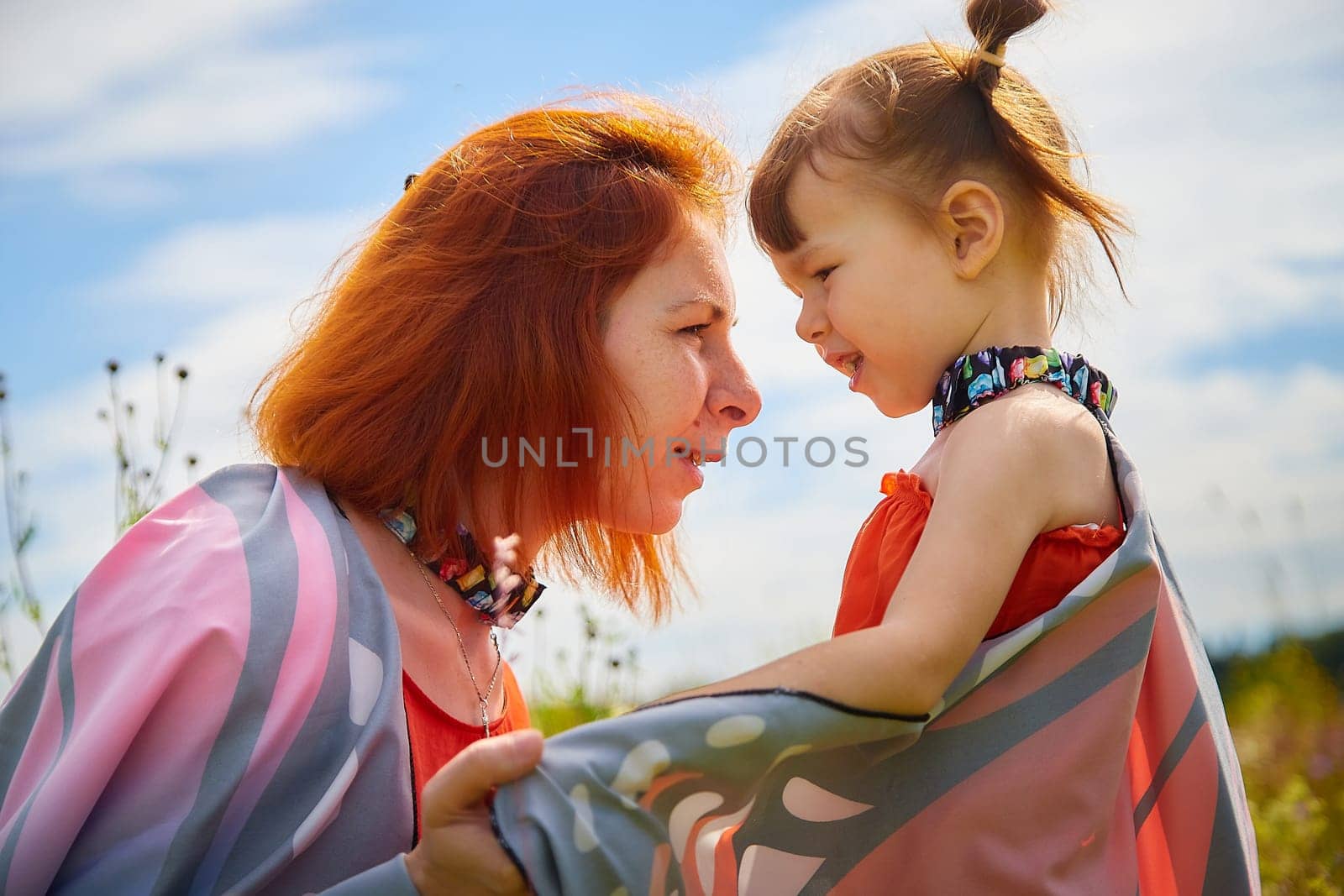 Happy female family with mother and daughter on green and yellow meadow full of grass and flower. Woman with red hair and blonde girl having fun, joy and hugs in sunny summer day. Concept family love by keleny