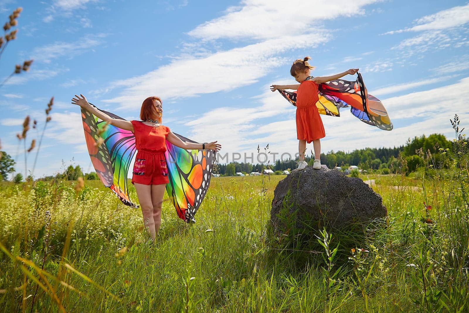 Happy female family with red haired mother and daughter with bright butterfly wings having fun on green and yellow meadow full of grass and flowers in sunny summer day. Concept family love