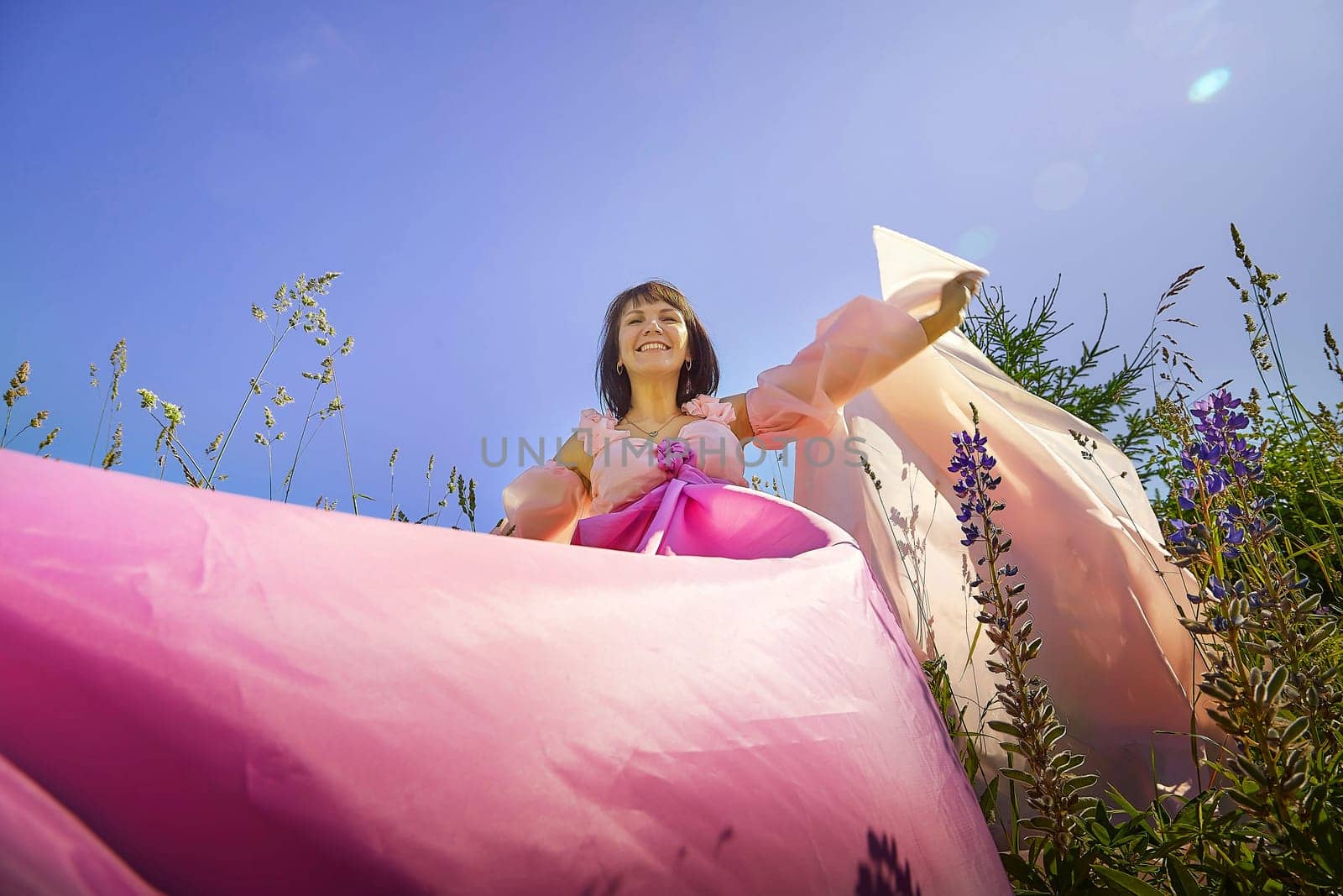 Beautiful girl in lush pink ball gown in green field during blooming of flowers and blue sky on background. Model posing on nature landscape as princess from fary tale