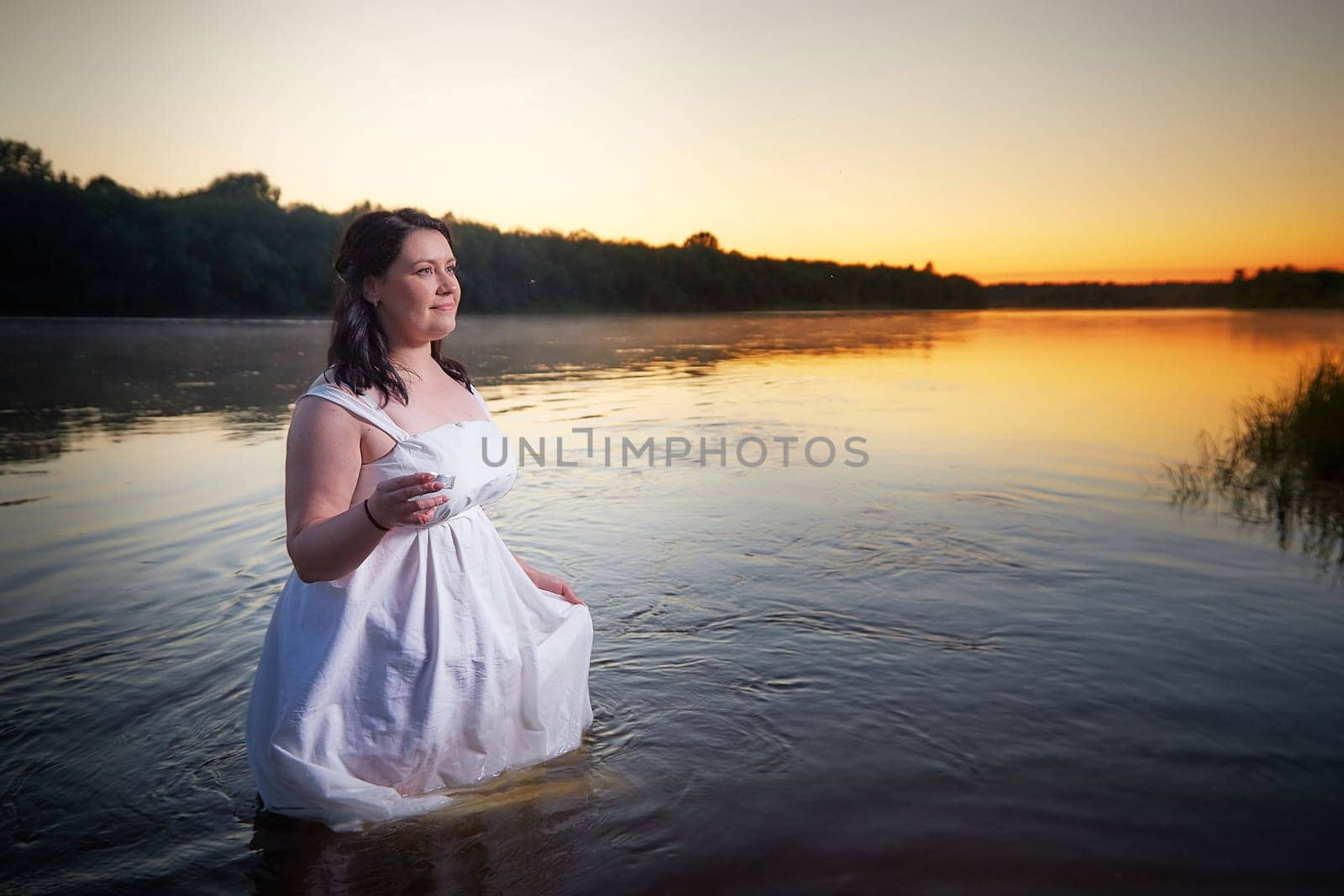 Slavic plump plump chubby girl in long white dress on the feast of Ivan Kupala with flowers and water in a river or lake on a summer evening by keleny