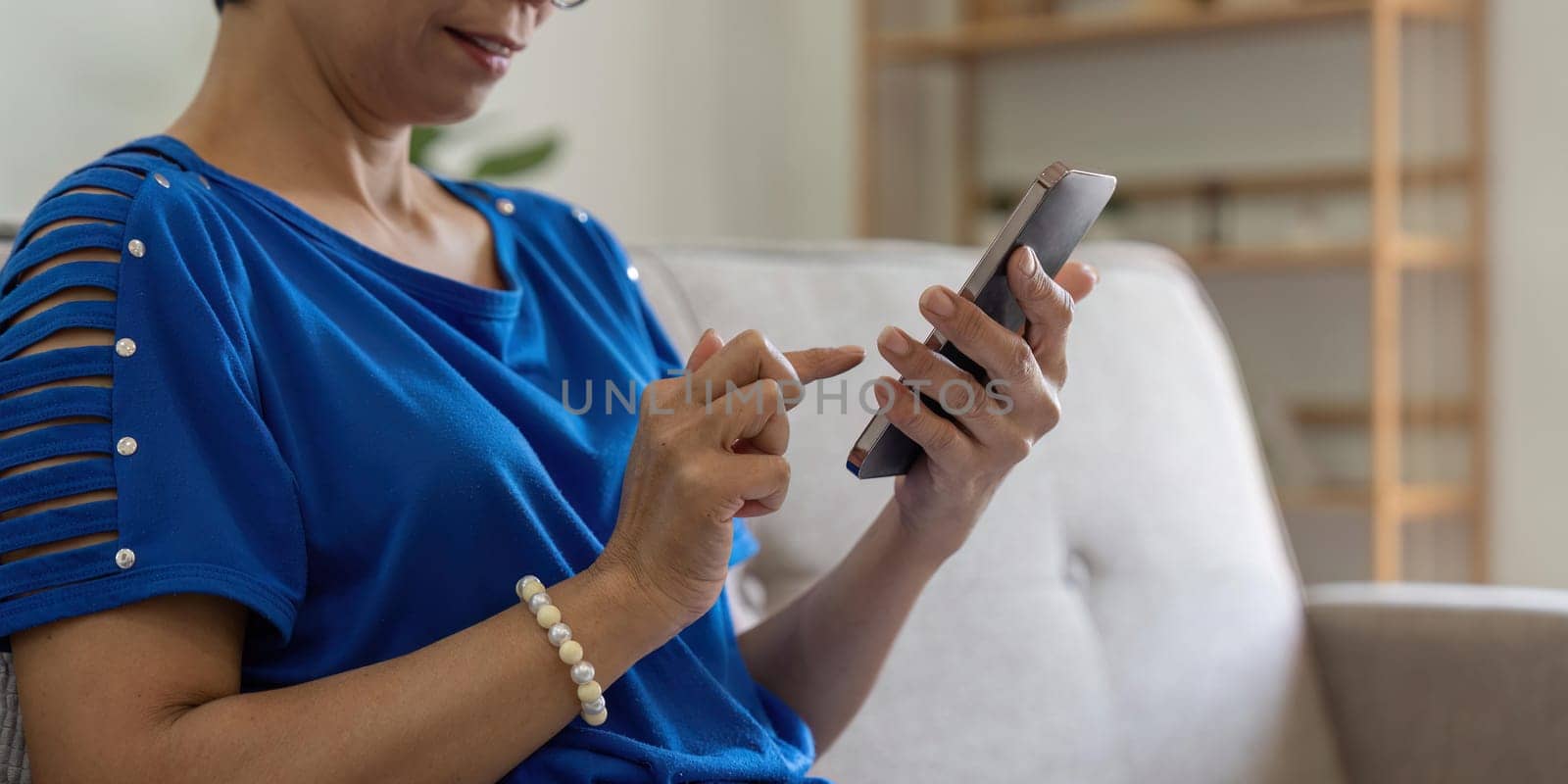 Smiling older asian woman sit rest on couch at home text or message online on modern cellphone gadget. Happy mature female relax browse internet on smartphone device. Elderly technology concept.