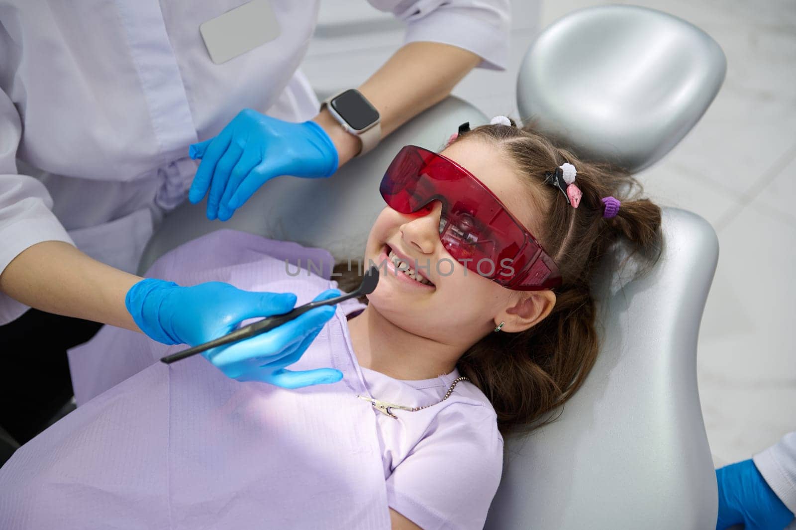 Cute little girl in dentist's chair, wearing UV protective glasses, smiling to a doctor dentist holding dental mirror while a teeth examination in pediatric dentistry clinic. Dental check-up concept