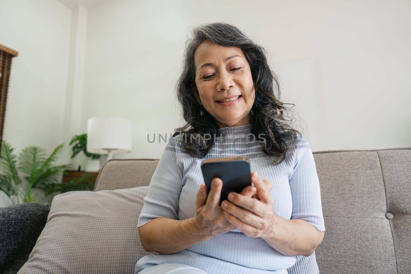 Smiling older asian woman sit rest on couch at home text or message online on modern cellphone gadget. Happy mature female relax browse internet on smartphone device. Elderly technology concept.