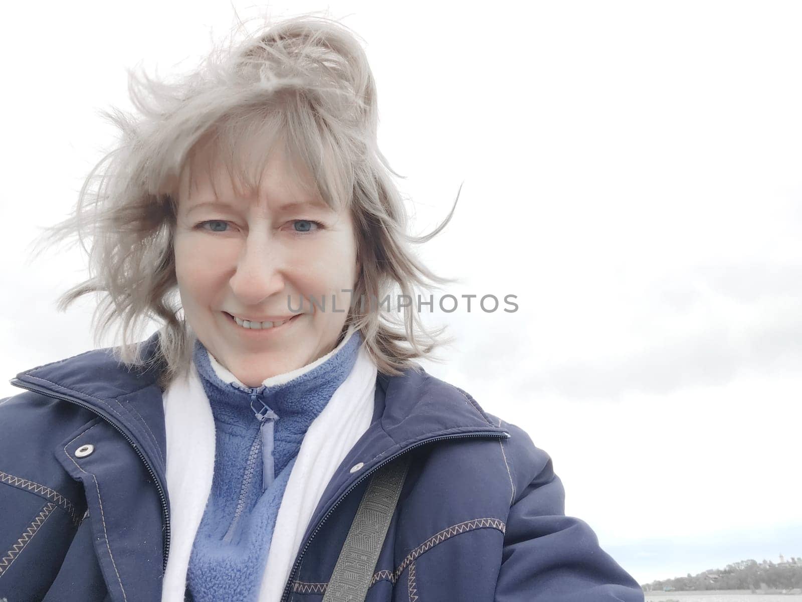 A blonde female tourist takes a selfie near the water of river or lake while traveling. The lovely girl or woman poses on her trip in summer, spring or autumn