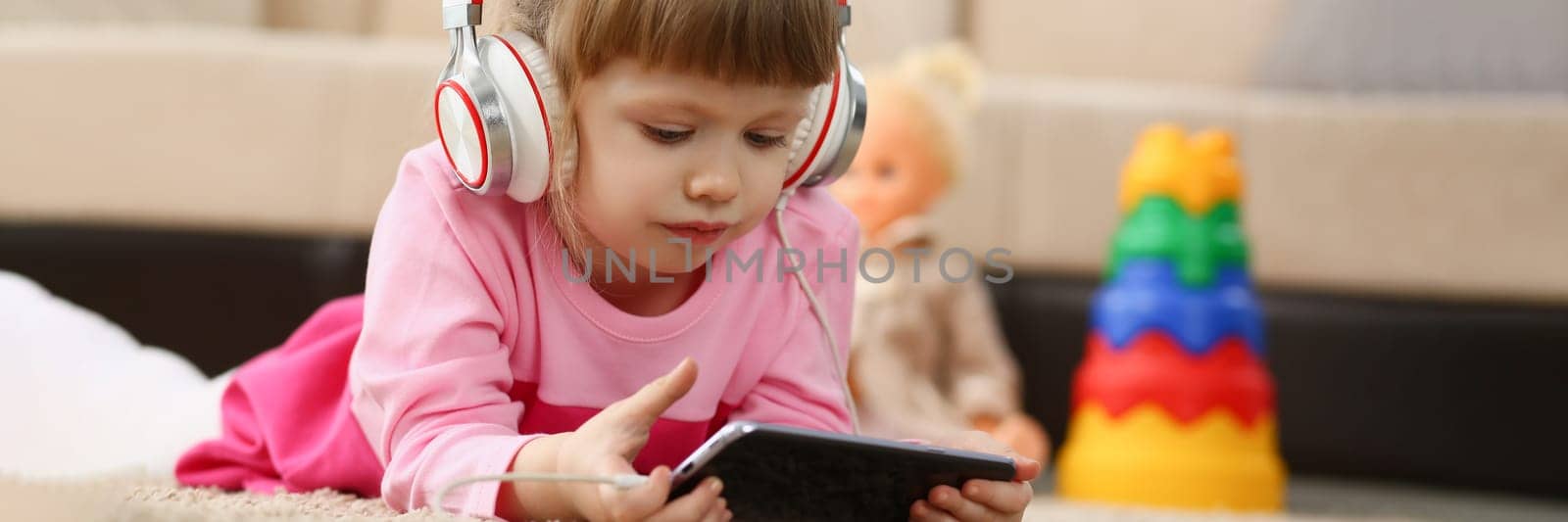 Little girl in headphones holds smartphone lying on floor. Children leisure watching cartoons and educational applications for children